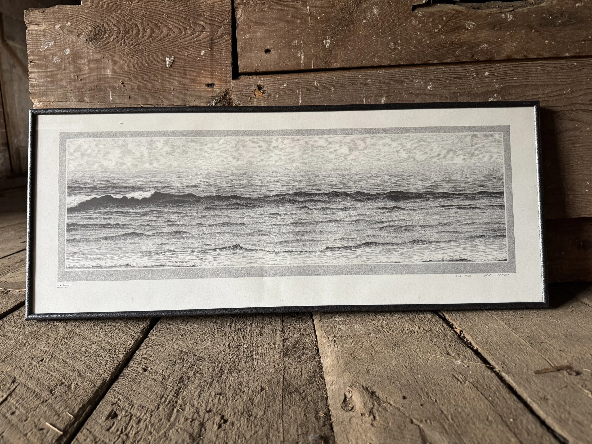 A black-and-white framed ocean photograph showing waves during the daytime, placed on a rustic wooden surface.