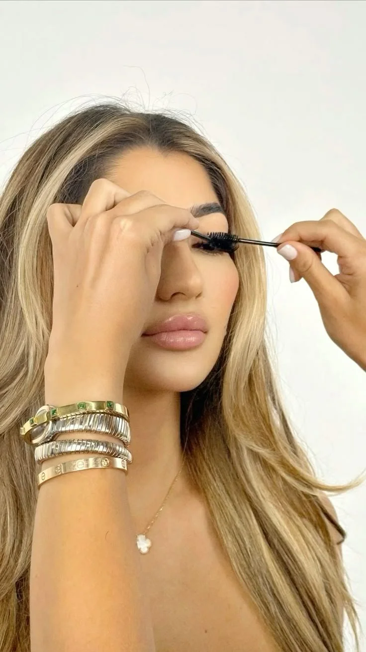 Woman having mascara applied to her eyelashes by another person, wearing bracelets and a necklace.