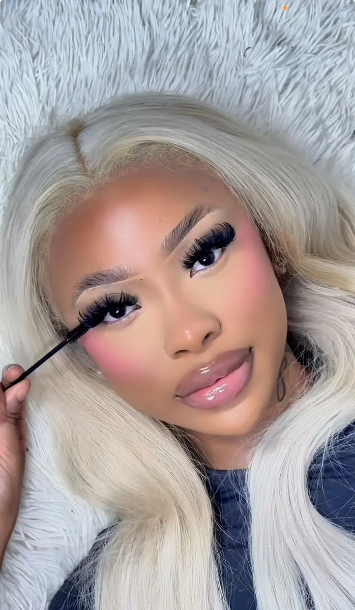 Woman with long blonde hair applying mascara, lying on a fluffy white surface.