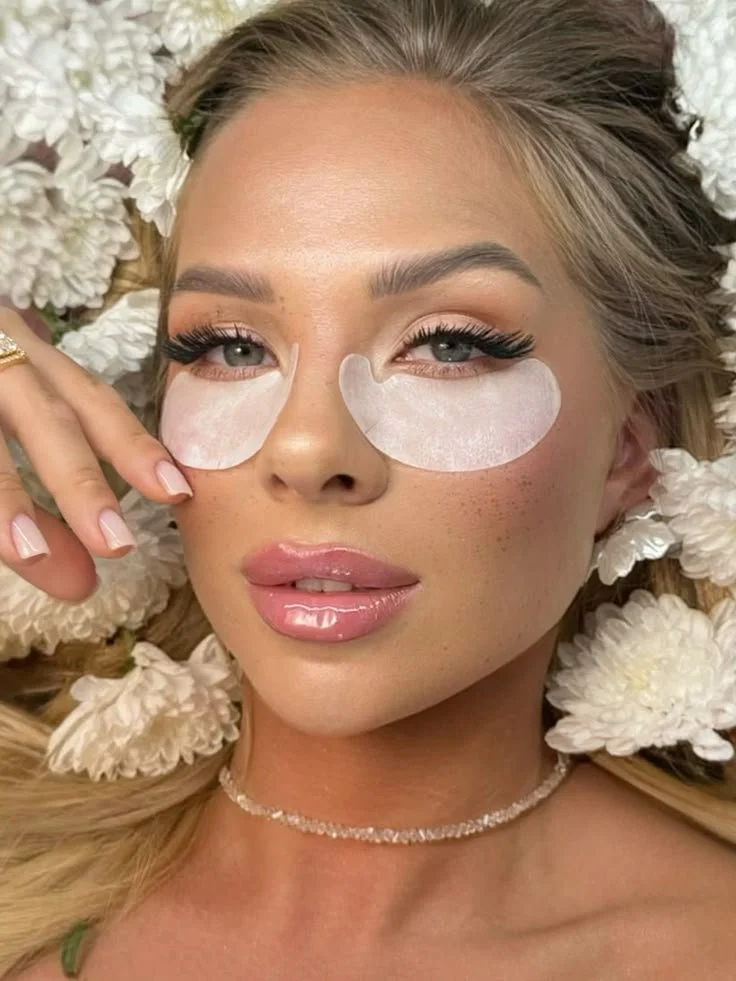 Close-up of a woman lying on white flowers, wearing under-eye patches, with styled hair and makeup.