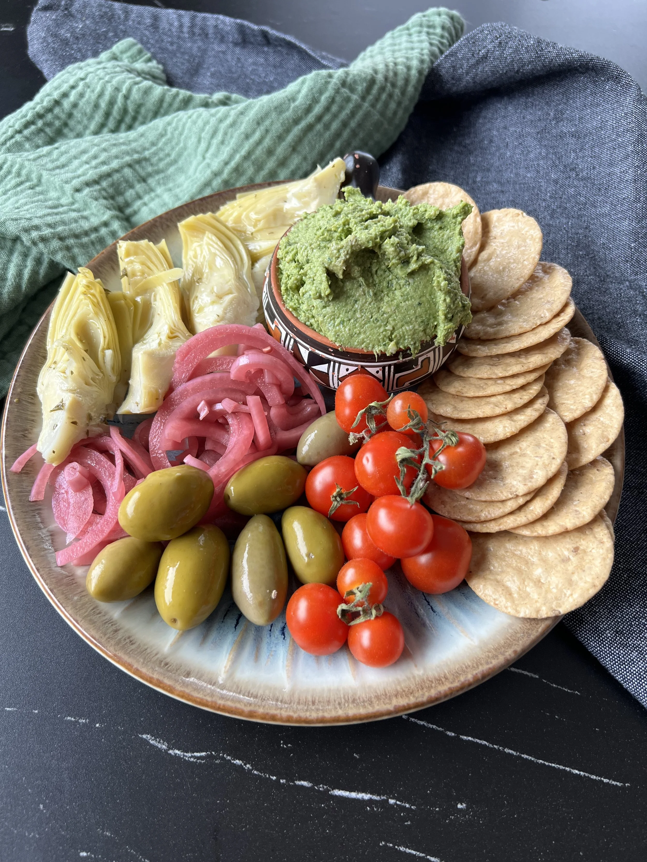 Dairy-free artichoke pesto served with crackers, marinated artichoke hearts, olives, pickled red onions, and cherry tomatoes on a platter.
