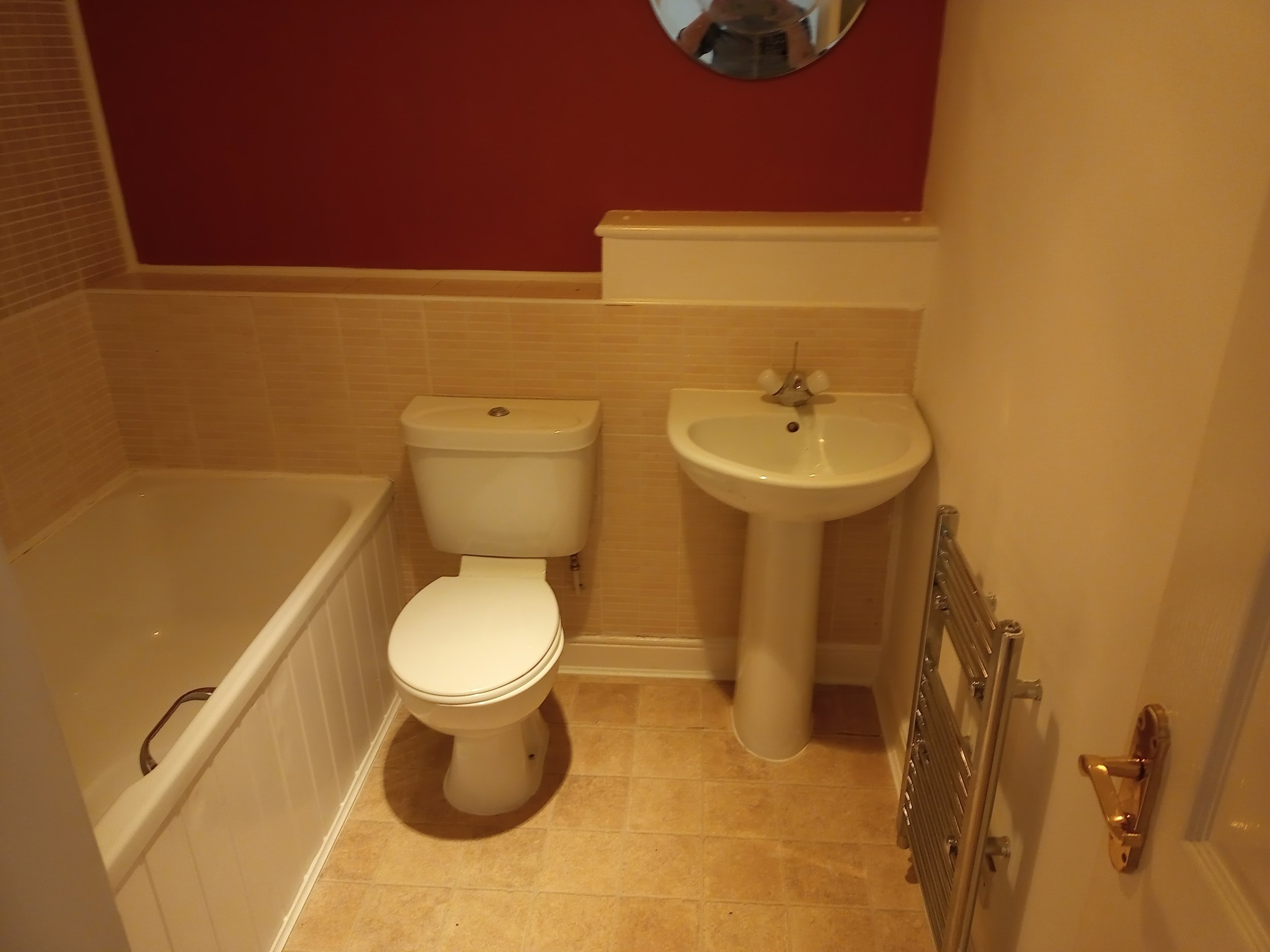 Bathroom with red accent wall, white bathtub, toilet, pedestal sink, round mirror, and towel radiator.