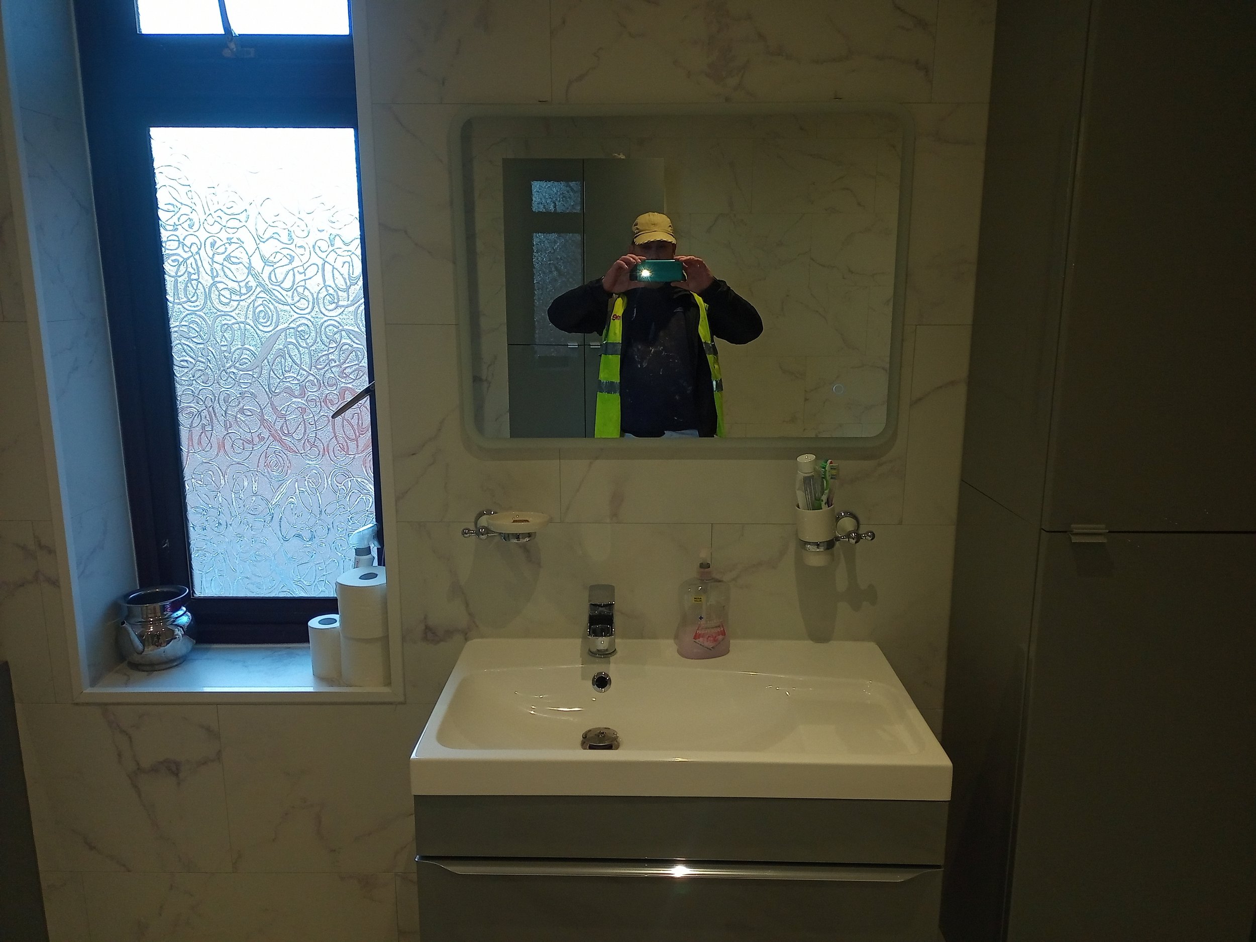 Modern bathroom with a wall-mounted sink, mirror, frosted glass window, soap dispenser, toothbrush holders, and decorative gray cabinetry.