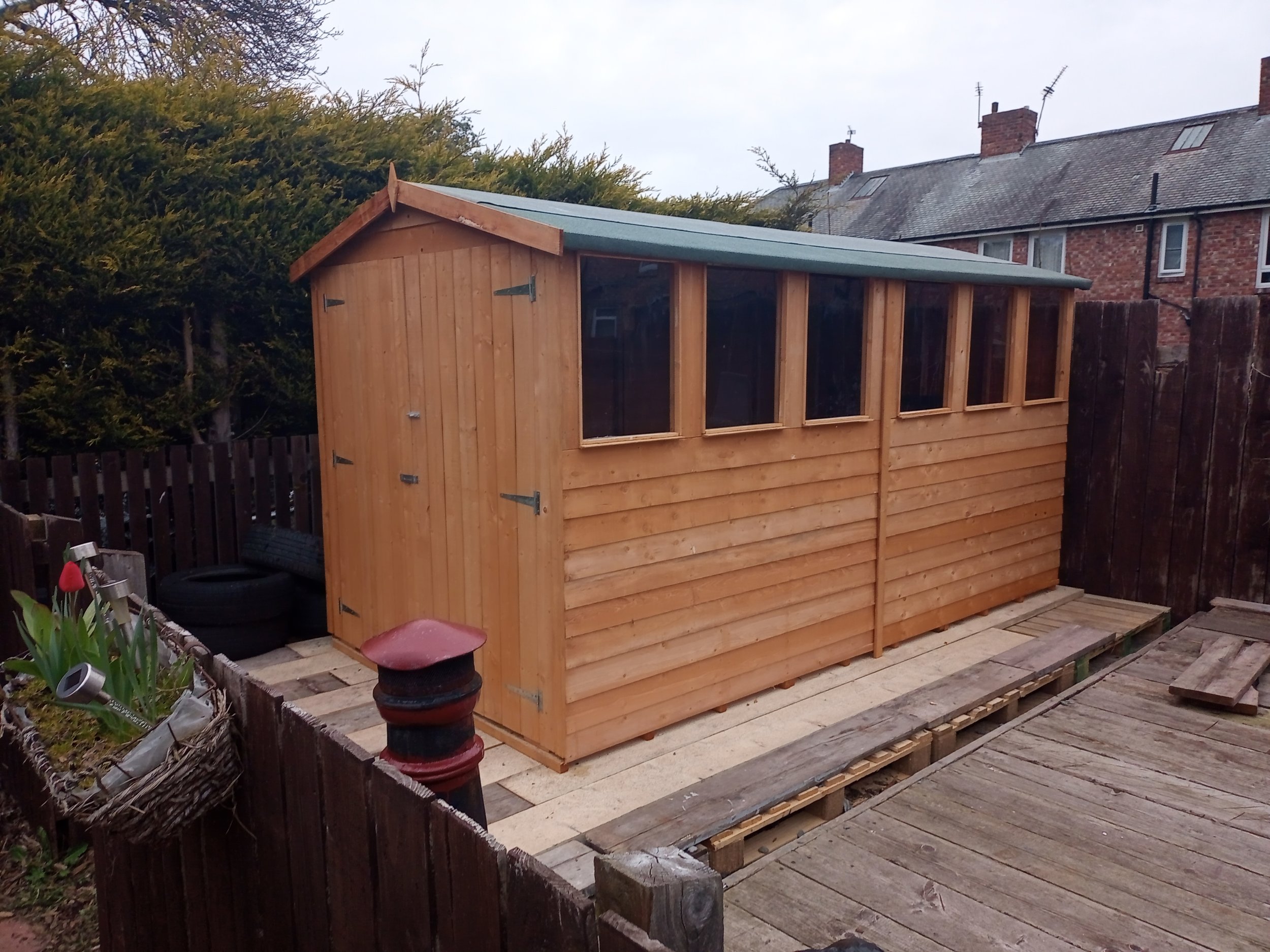 Wooden shed with windows in a fenced backyard, surrounded by plants and a wooden deck.