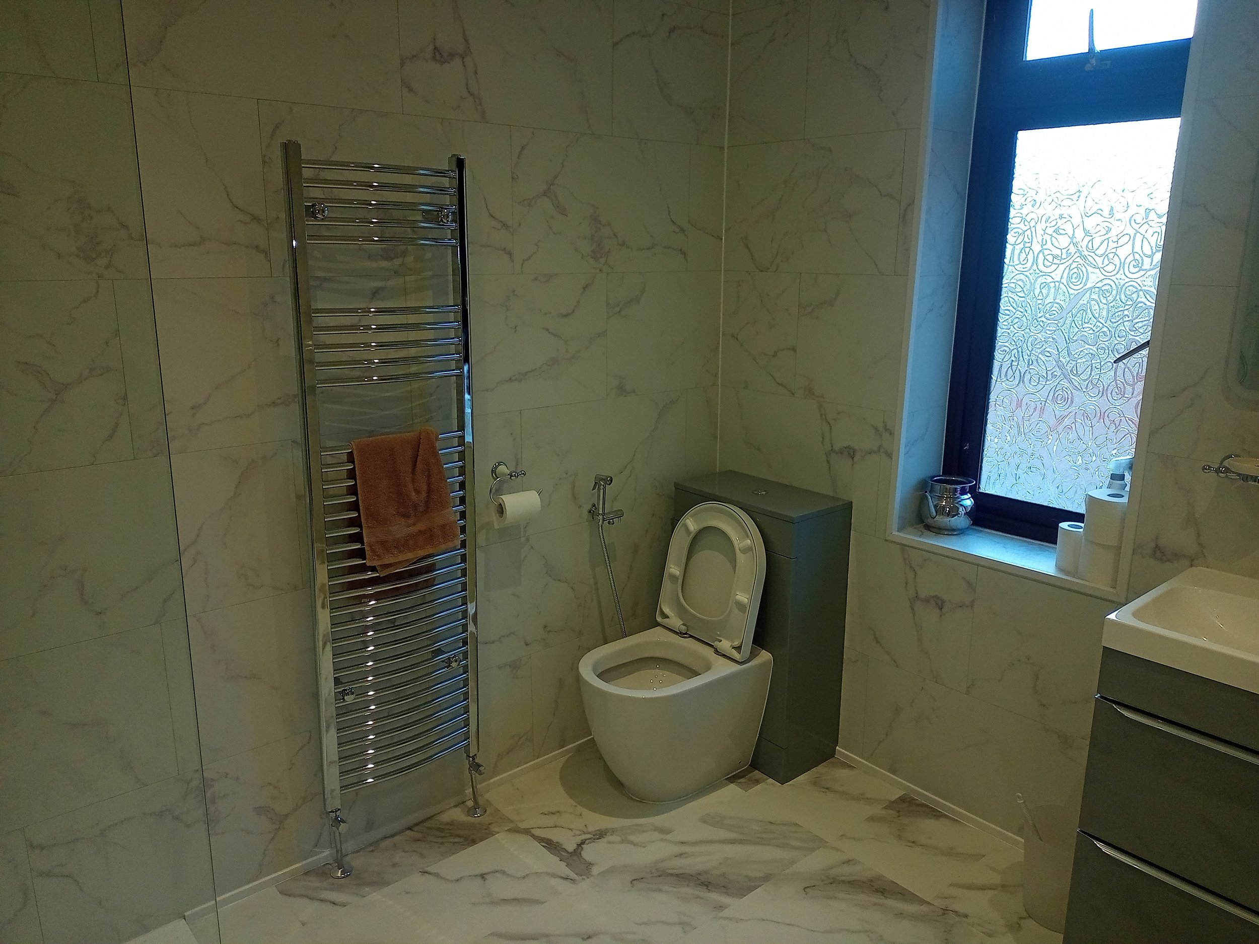Modern bathroom with marble tiles, featuring a wall-mounted toilet, stainless steel towel warmer, frosted window with decor, and a sleek vanity with a sink.