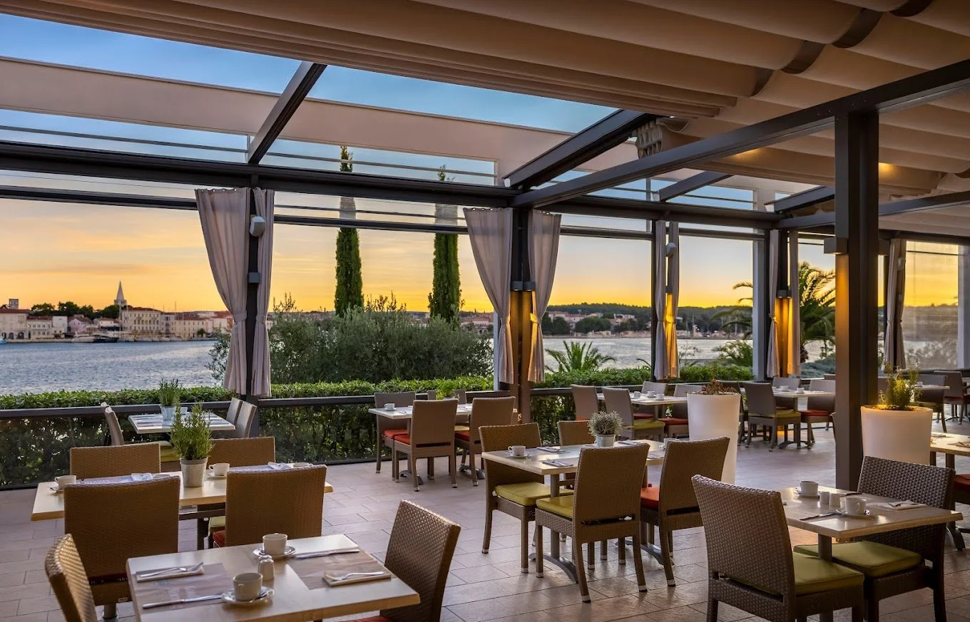 Outdoor restaurant patio with tables and chairs overlooking a waterfront at sunset, framed by greenery and glass walls.