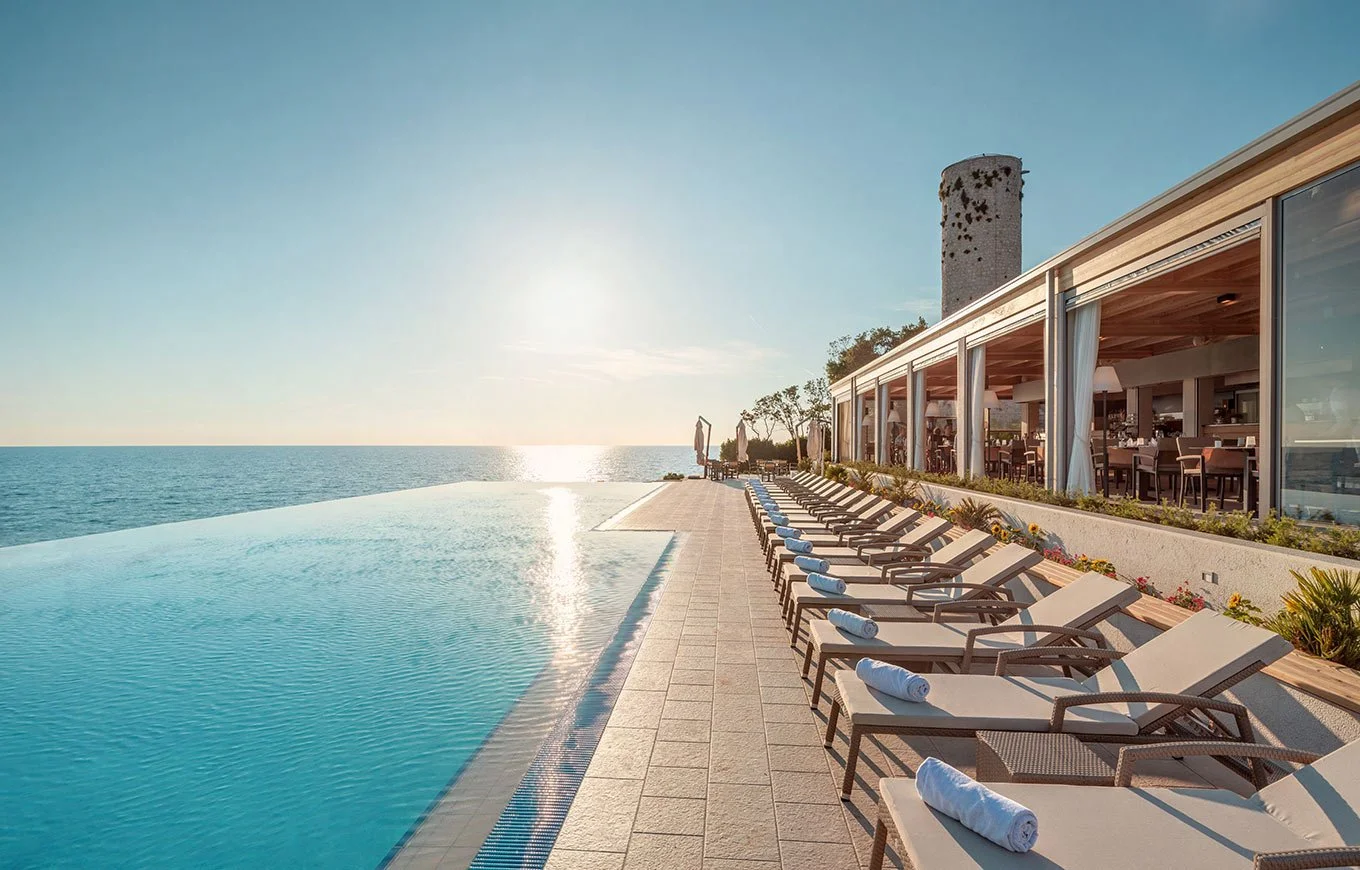 Infinity pool with lounge chairs, ocean view at sunset, resort building with outdoor seating.