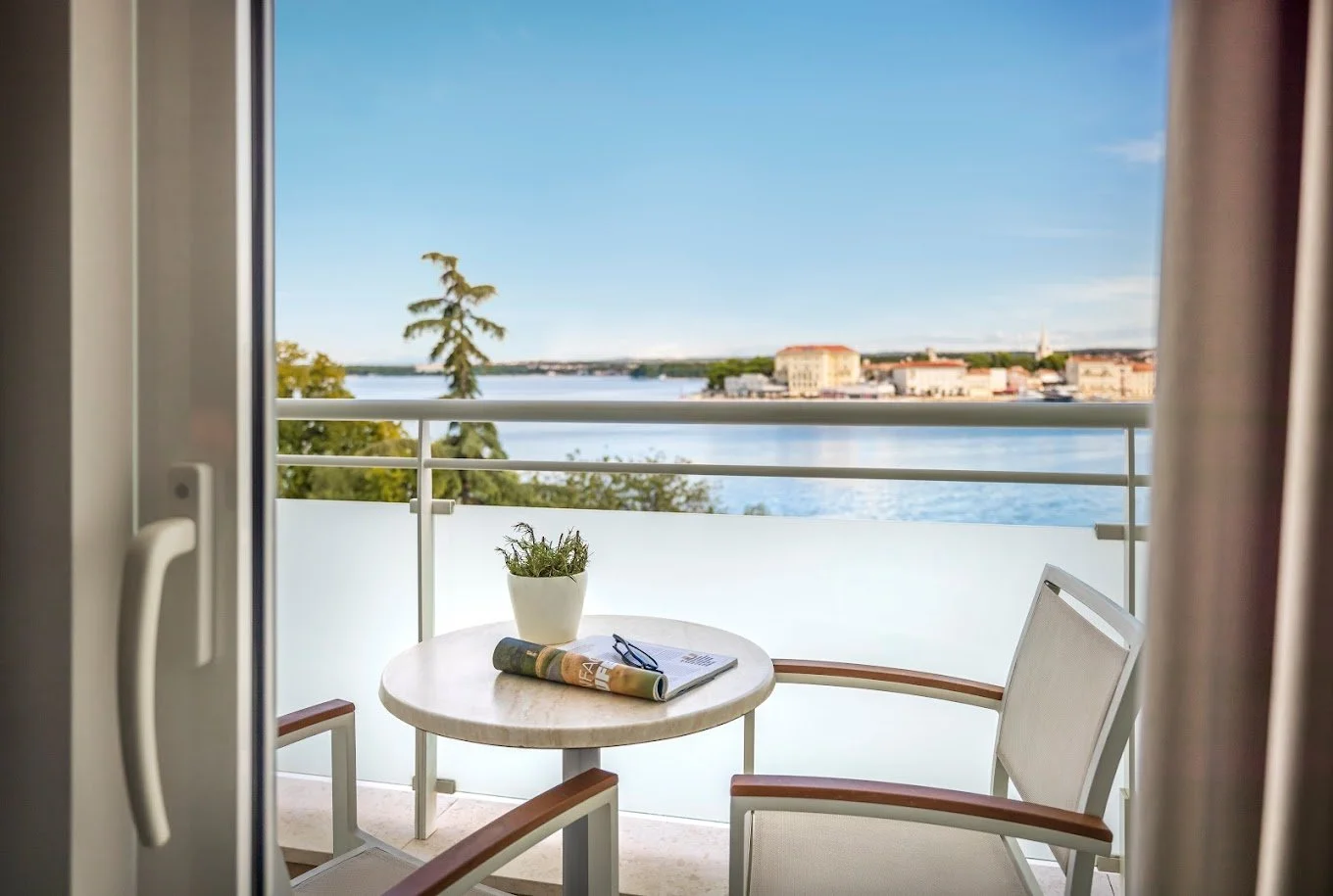 View from a balcony overlooking water, with a round table and two chairs, a potted plant, and a rolled magazine or brochure. Buildings and trees are visible in the background under a clear blue sky.