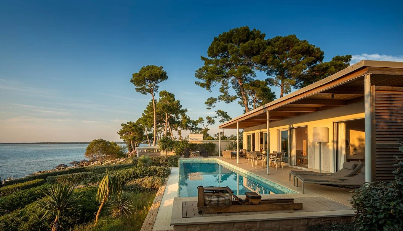 Modern house with a pool overlooking a waterfront view, surrounded by trees and a garden, under a clear blue sky.