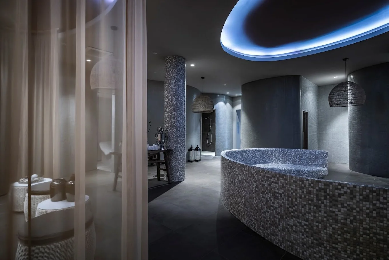 Modern spa interior with curved walls, mosaic tiles, and soft lighting. Features include a seating area, tables with decor, hanging lamps, and shower area.