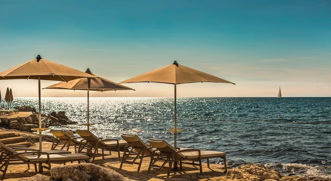 Beach scene with lounge chairs and umbrellas by the ocean, sailboat on horizon.