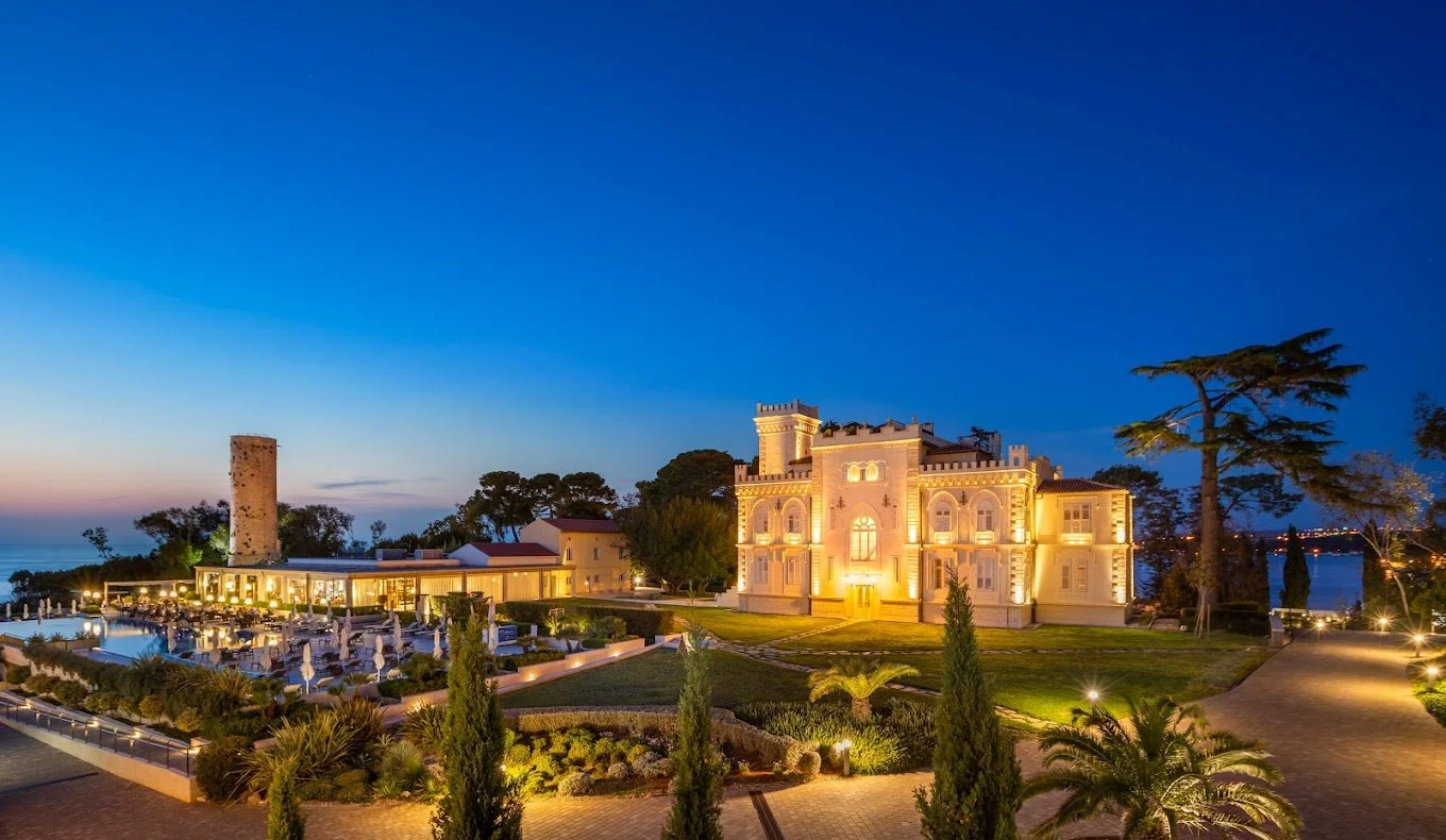 Elegant villa and tower with outdoor terrace lit at dusk near a body of water.