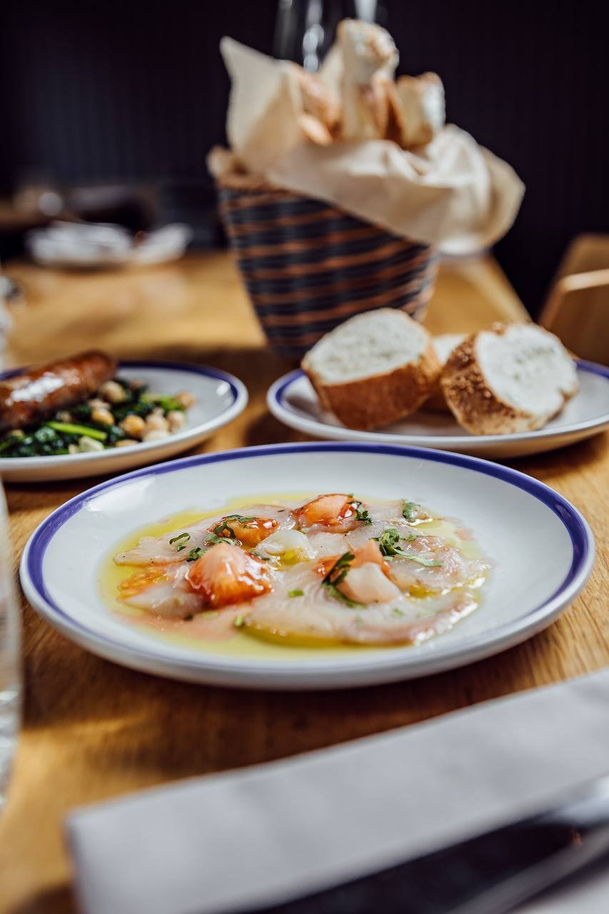 Plate of seafood ceviche with olive oil and herbs, slices of bread, basket with bread rolls, and a sausage dish on a wooden table.