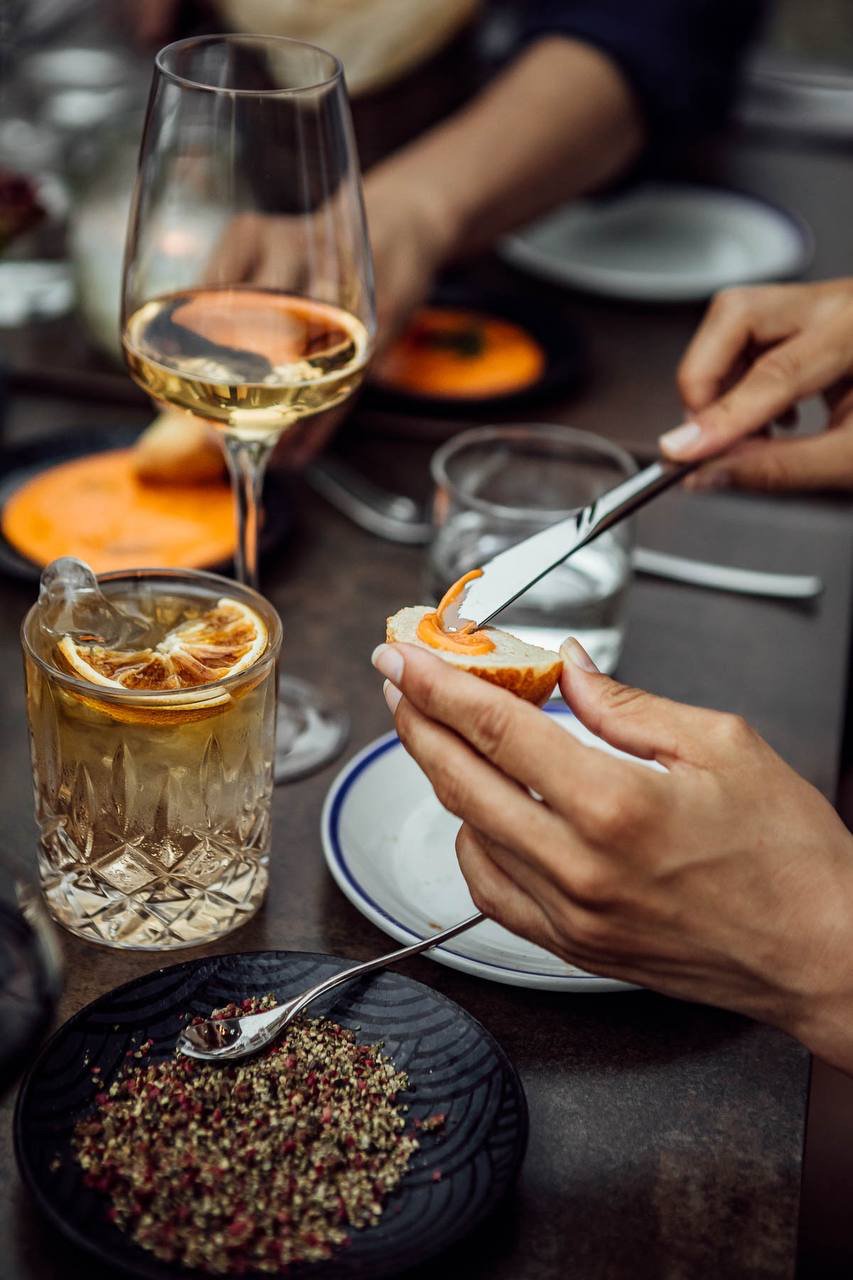 Person spreading topping on bread with a drink and seasonings in the foreground.