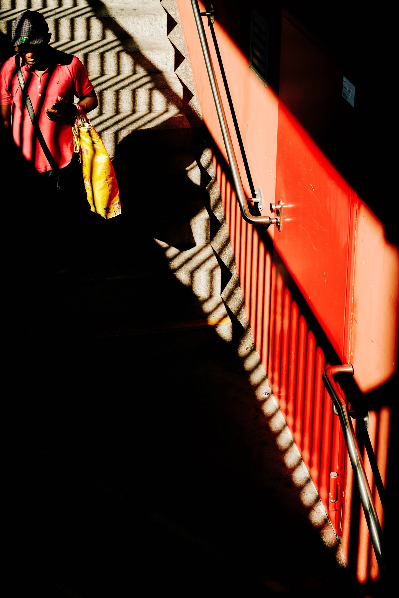 A person in a red shirt and baseball cap walking down concrete stairs, carrying a yellow shopping bag, with shadows of the stair railing cast onto the wall and stairs.