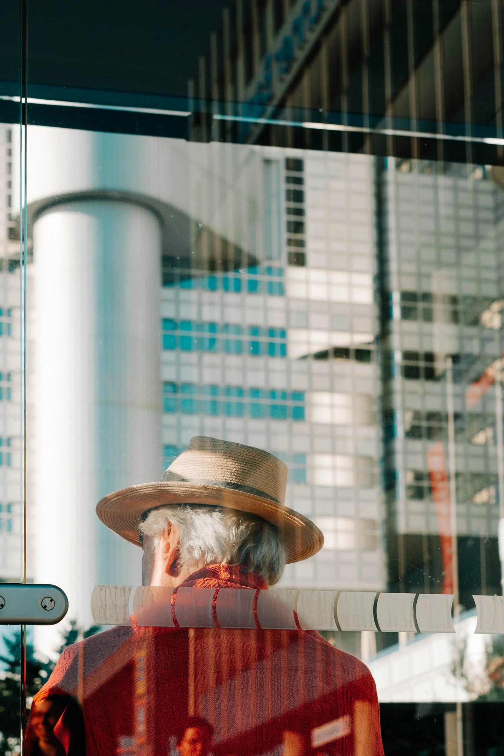 Old woman with gray hair wearing a wide-brimmed straw hat and red coat, viewed from behind, siting inside a glass building with cityscape reflections of tall modern office buildings.