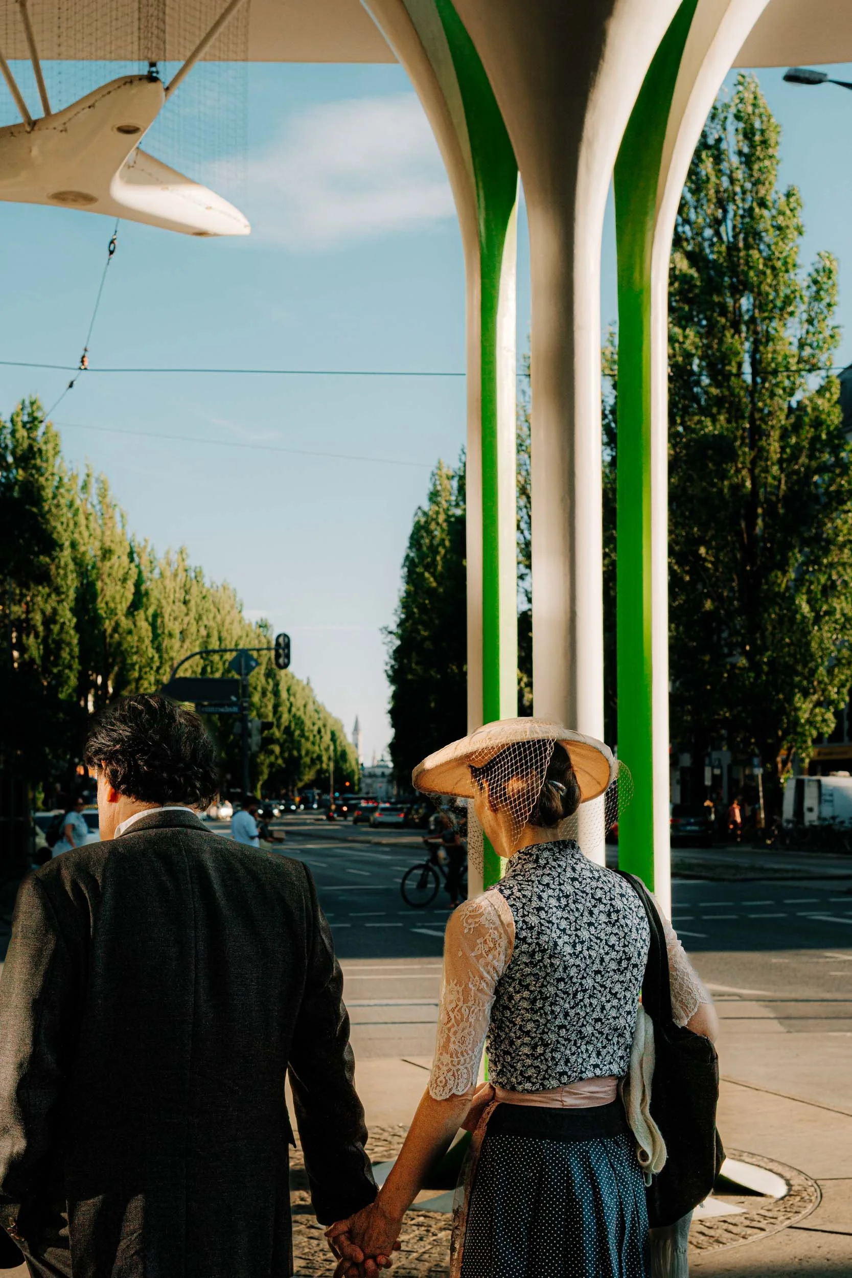 A couple holding hands at a city crosswalk on a sunny day with trees and traffic in the background. The woman wears a vintage hat with a veil and lace top, and the man wears a dark blazer.
