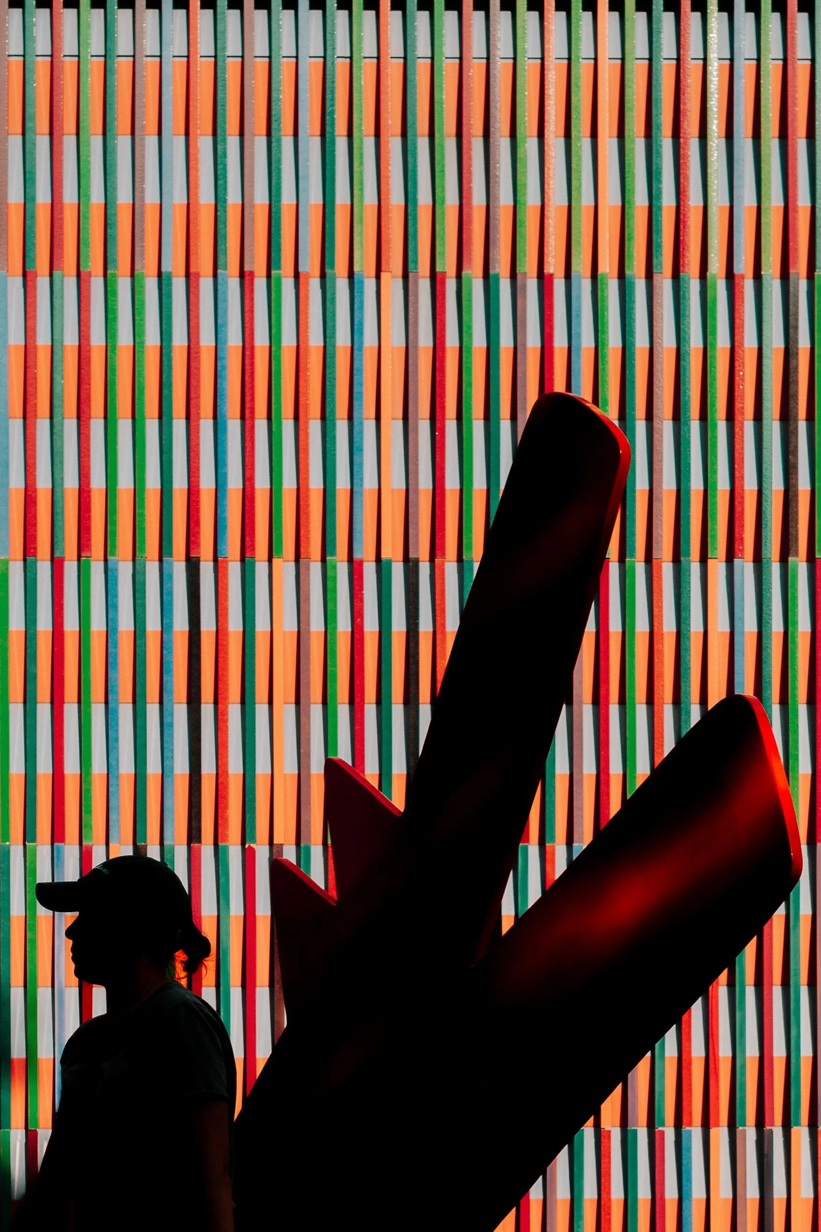 Silhouette of a person wearing a baseball cap walking past a large, stylized red arrow pointing upward against a colorful, geometric patterned wall.