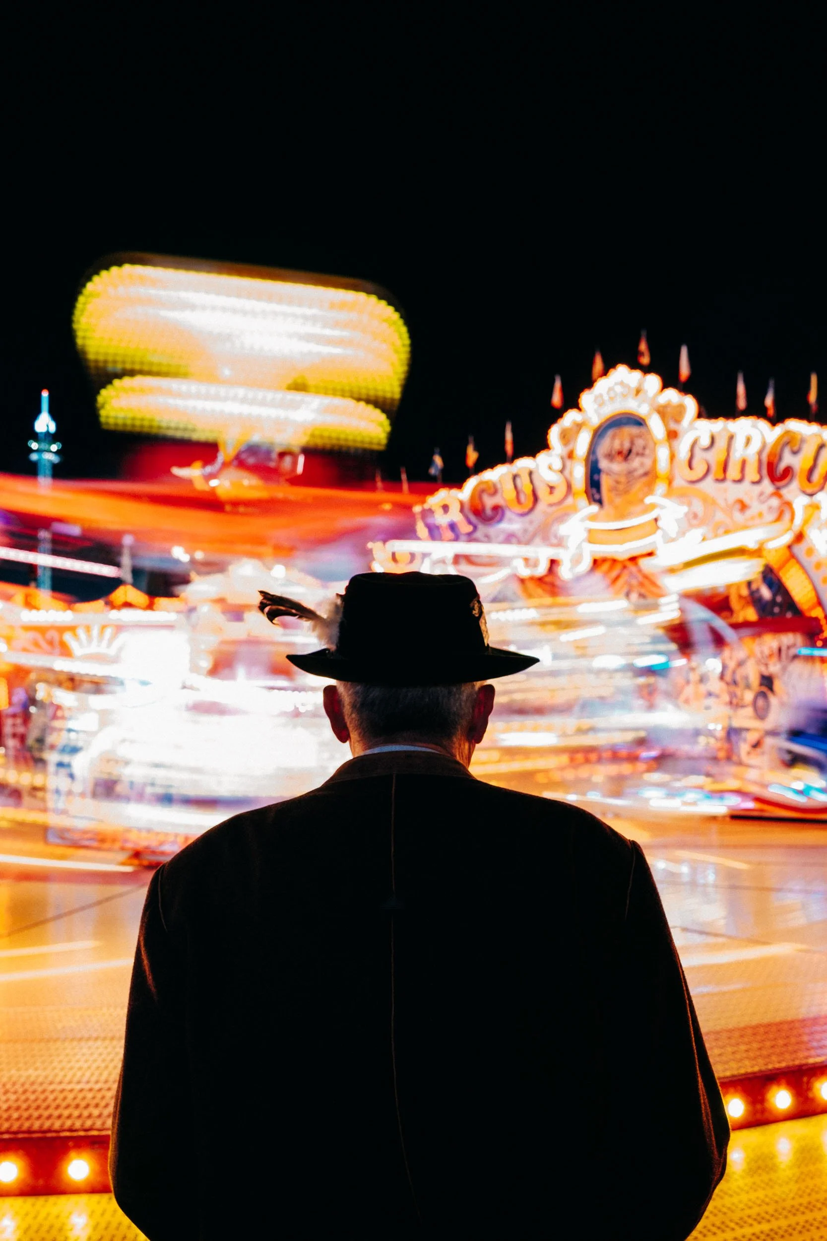 A man wearing a black hat with a feather stands in front of a brightly lit carnival ride at night, with blurred lights and motion indicating spinning attractions.