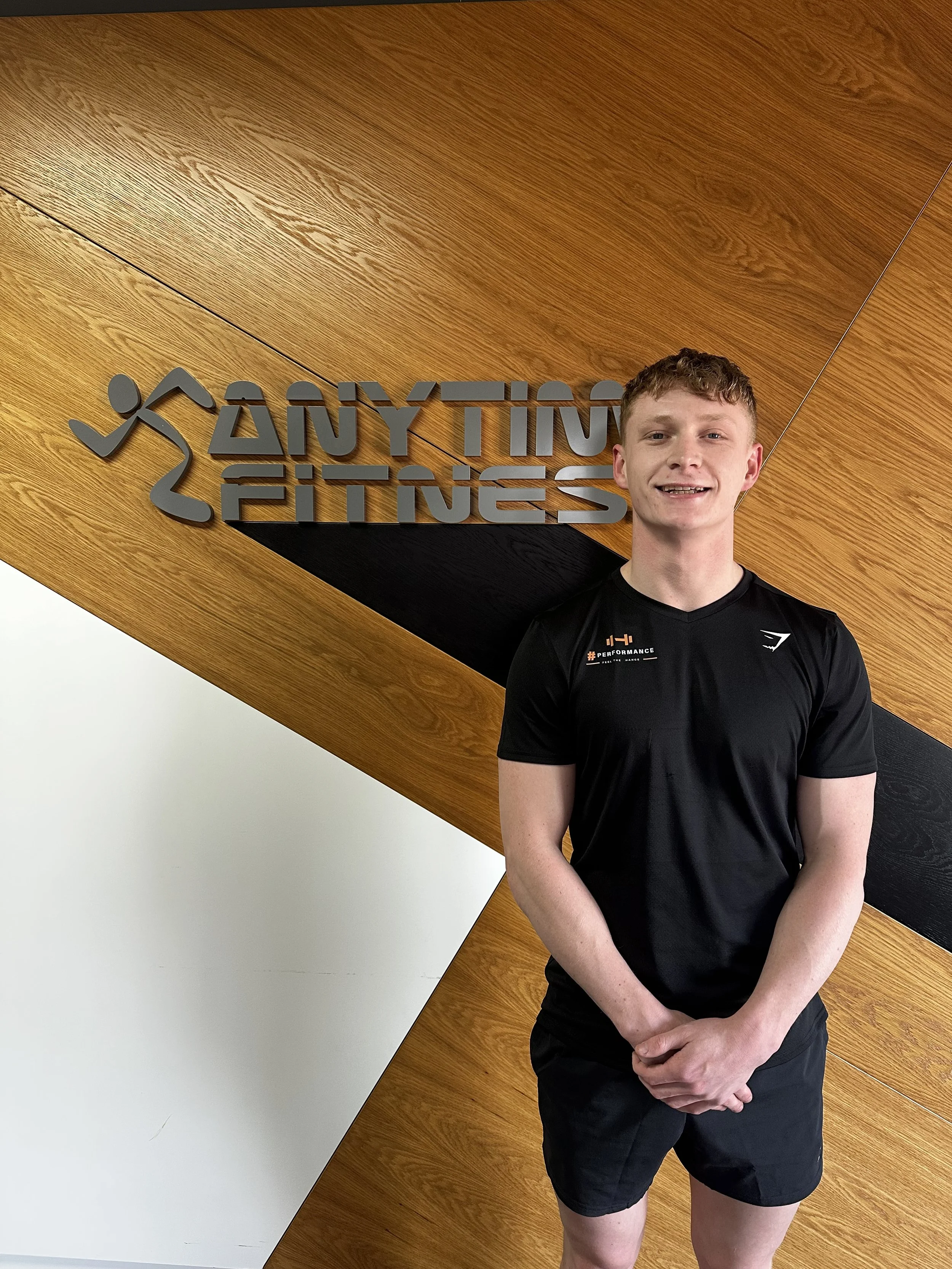 A man standing indoors in front of a wooden wall with the text 'ANY TIME FITNESS' logo. He is wearing a black athletic shirt and shorts, smiling at the camera.