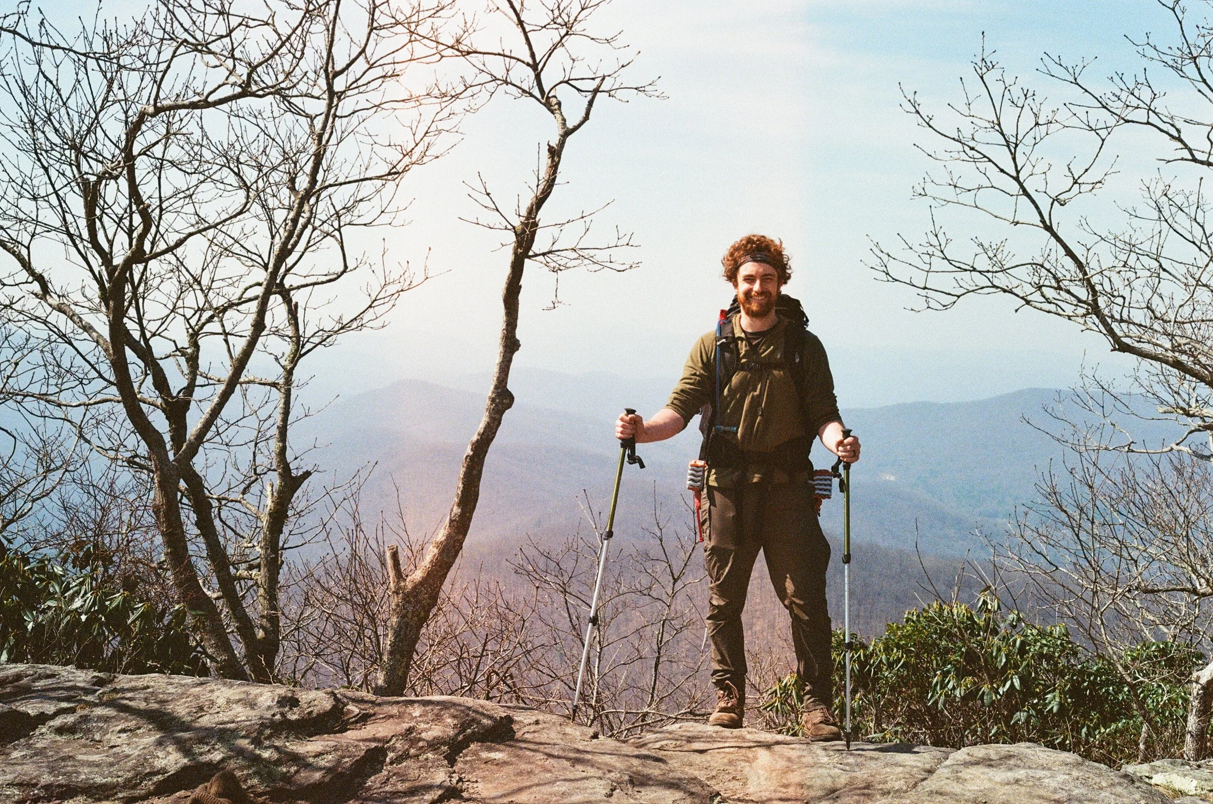 A person with hiking gear stands on a rocky surface holding trekking poles, surrounded by bare trees, with a mountainous landscape in the background.