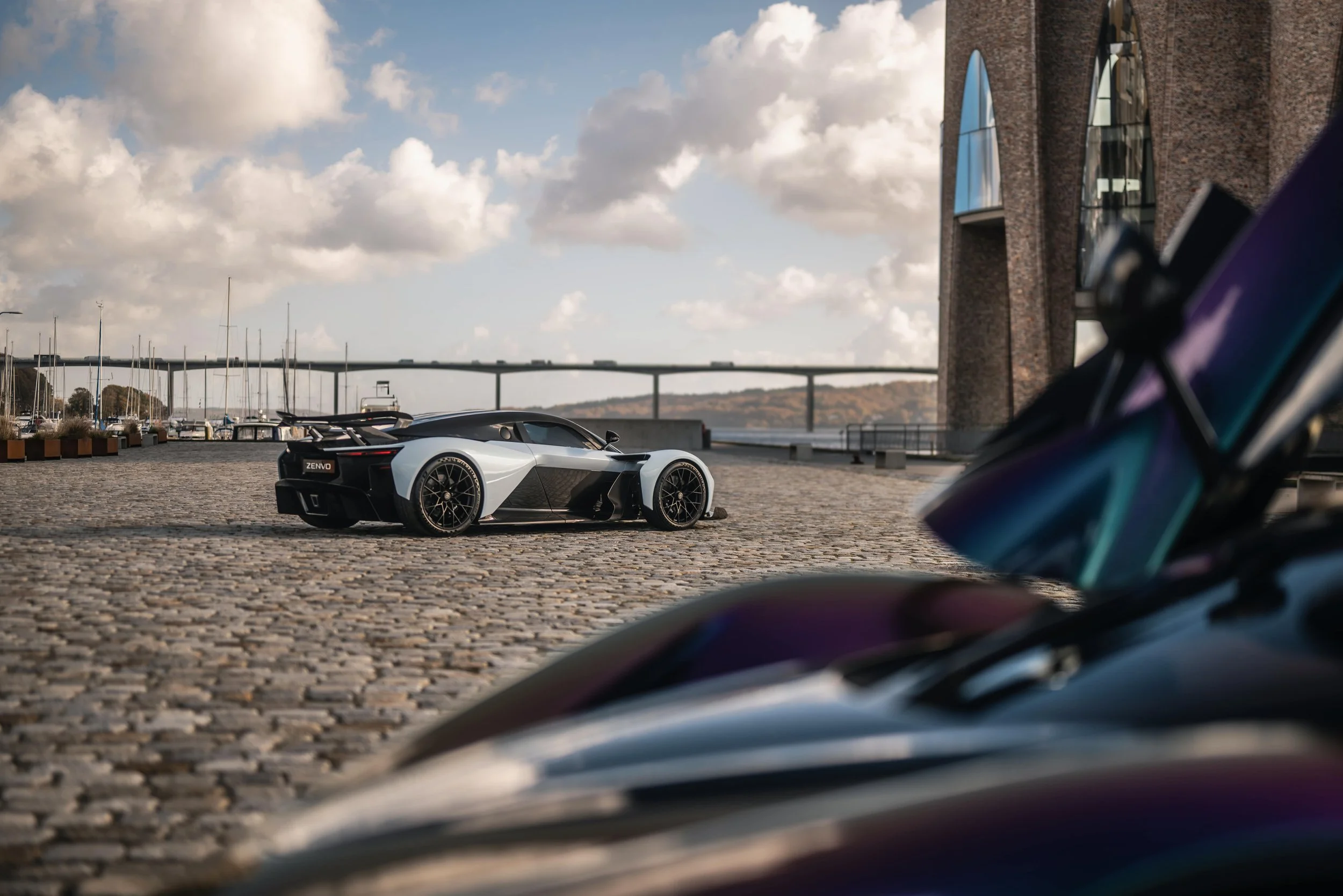 Zenvo Aurora Mjølner Hypercar in zweifarbiger Lackierung, geparkt auf Kopfsteinpflaster mit Yachthafen und Brücke im Hintergrund.