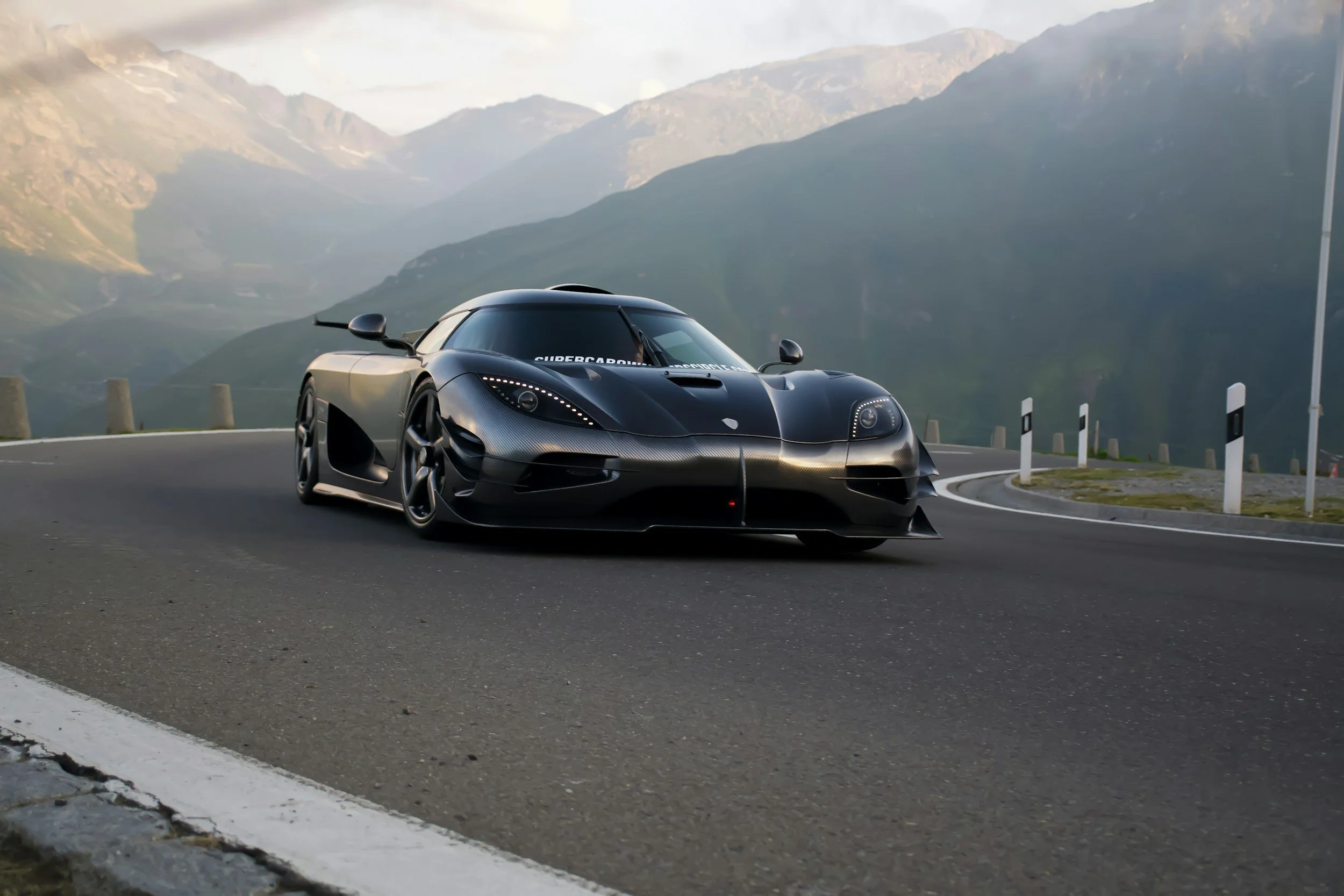Ein schwarzes Hypercar, Koenigsegg Jesko Absolut fährt auf einer kurvigen Bergstraße mit Bergen im Hintergrund.