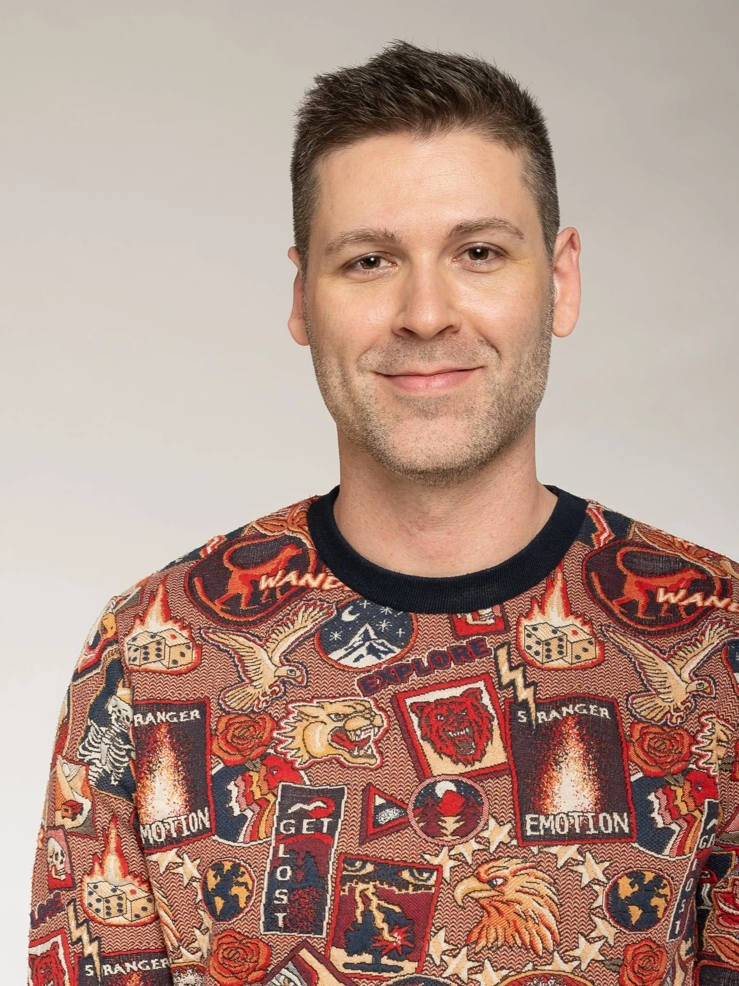 Mann mit kurzen dunklen Haaren, leichtem Bart, trägt ein buntes, patchworkartiges Pullover mit Tiermotiven und Schriftzügen, vor hellem Hintergrund.