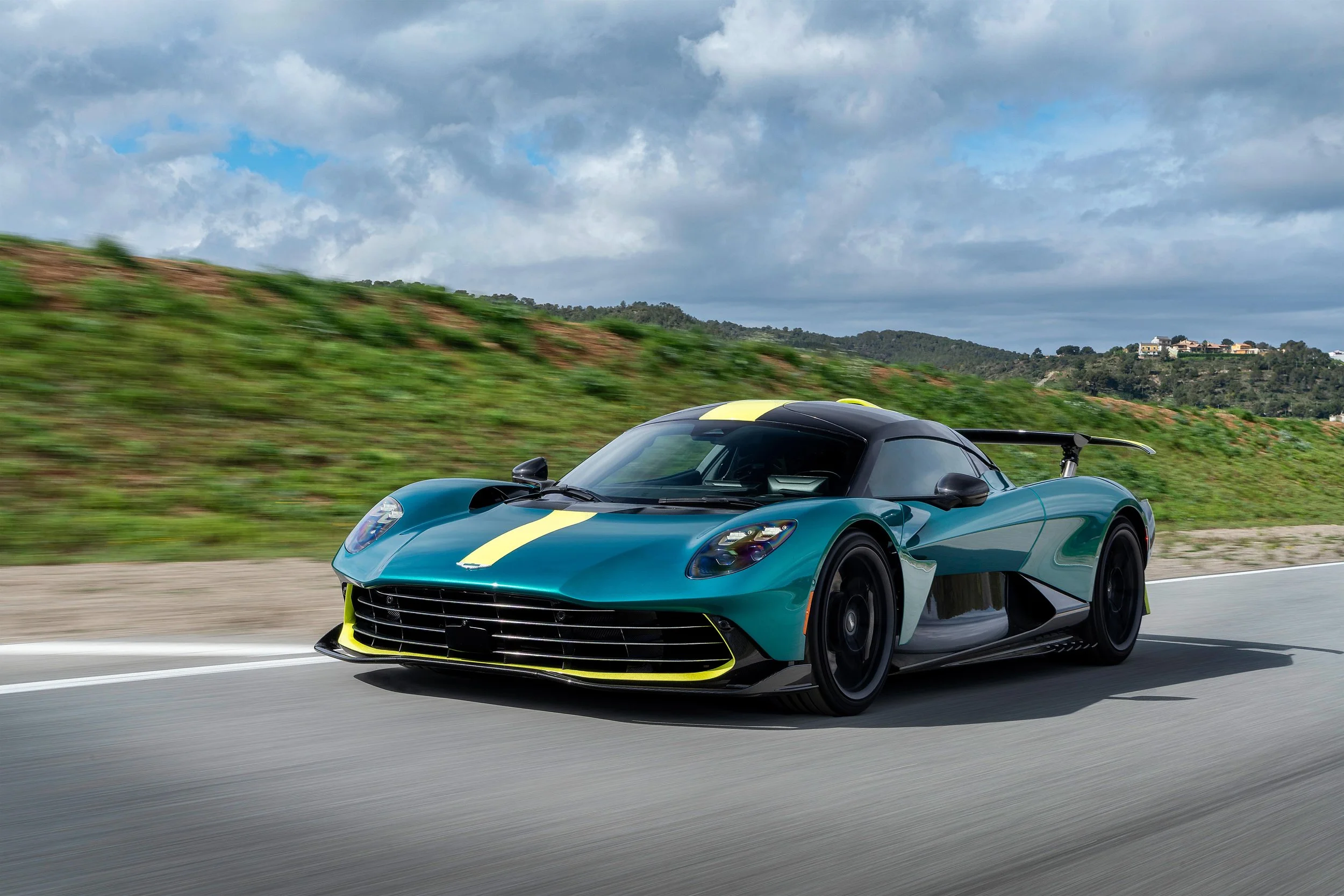 Aston Martin Valhalla in Racing Green mit gelbem Mittelstreifen, aufgenommen in Bewegung auf einer Landstraße während einer Testfahrt, Seitenansicht vorne links.
