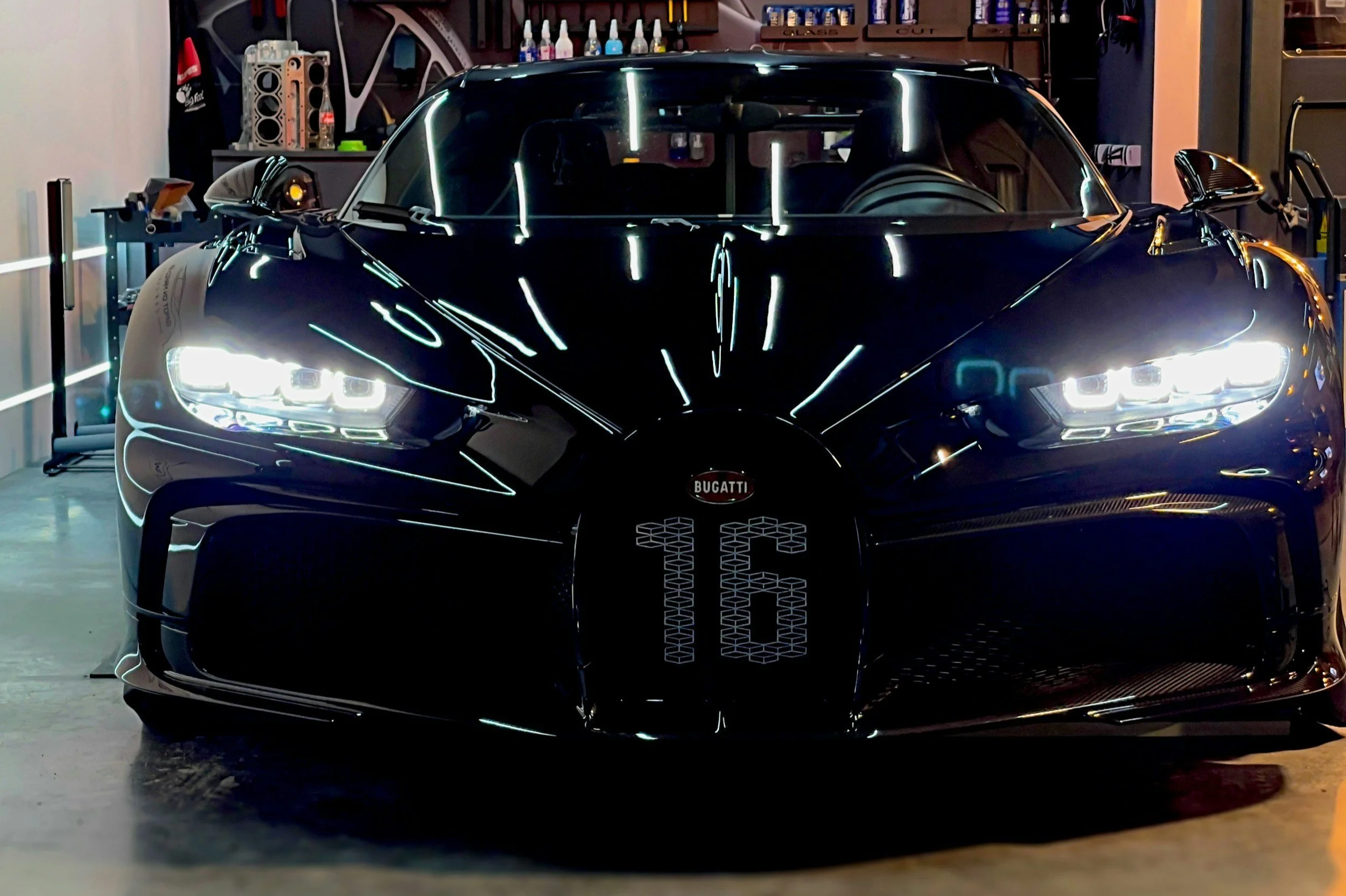 Schwarzer Bugatti Chiron in einer Garage, reflektierende Oberfläche, Frontansicht mit leuchtenden Scheinwerfern und Bugatti-Logo, Nummer 16 auf der Motorhaube.