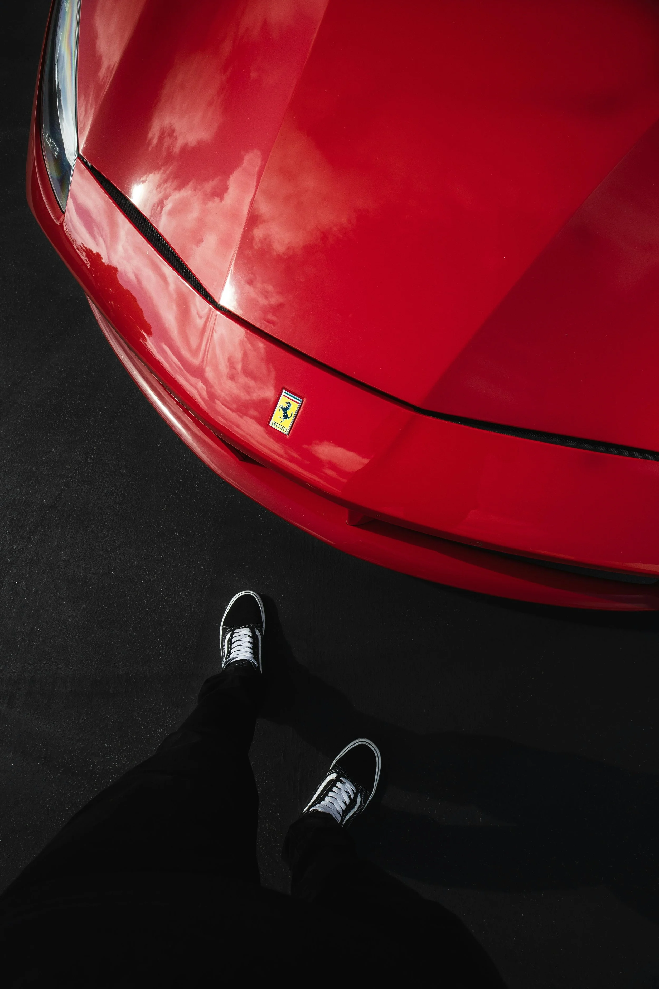 Vogelperspektive auf eine rote Ferrari-Sportwagenfront mit Ferrari-Logo, schwarze Schuhe mit weißen Schnürsenkeln, schwarzer Boden, Himmelreflexion auf Karosserie.