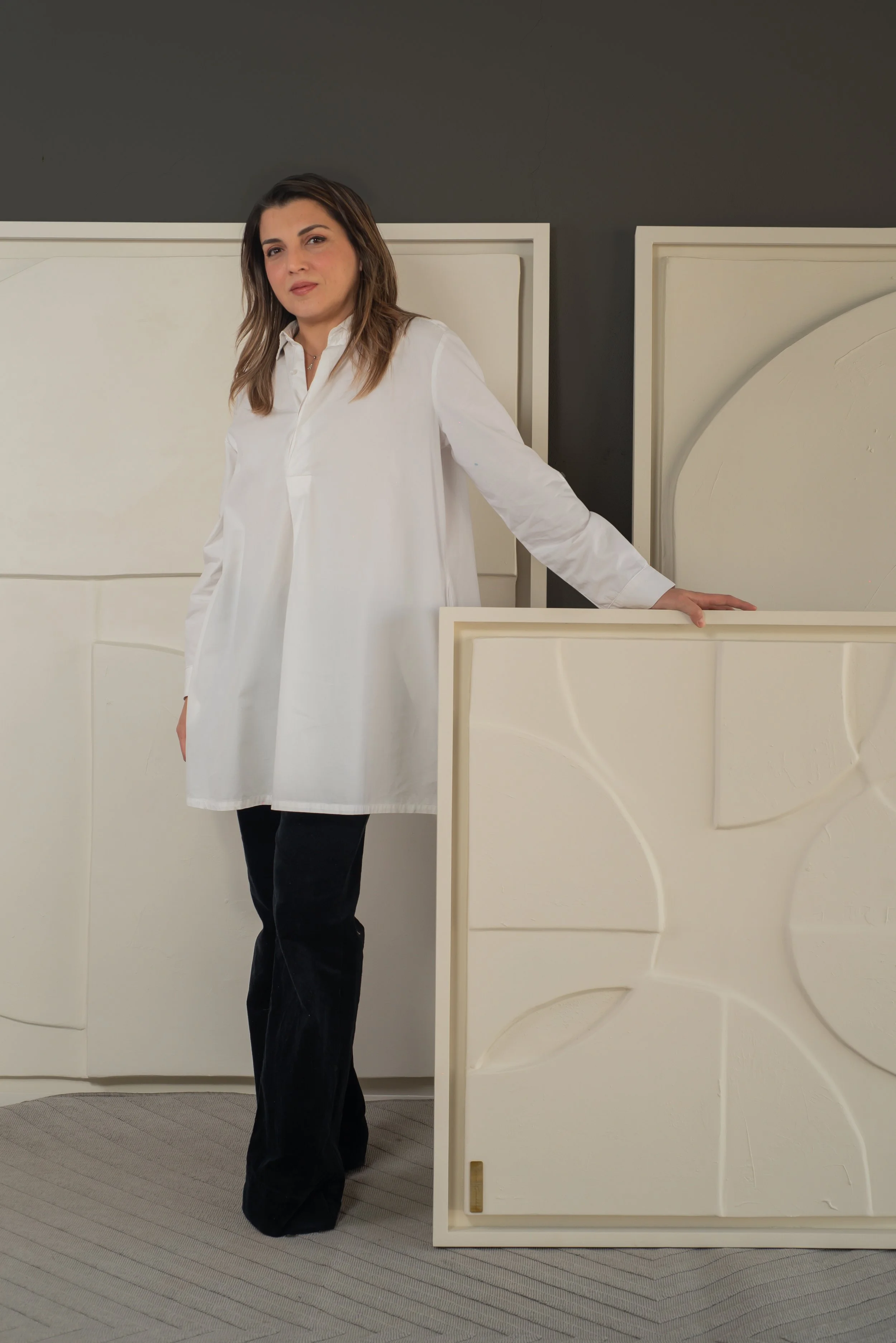 Mujer con blusa blanca y pantalones oscuros posando junto a paneles decorativos blancos.