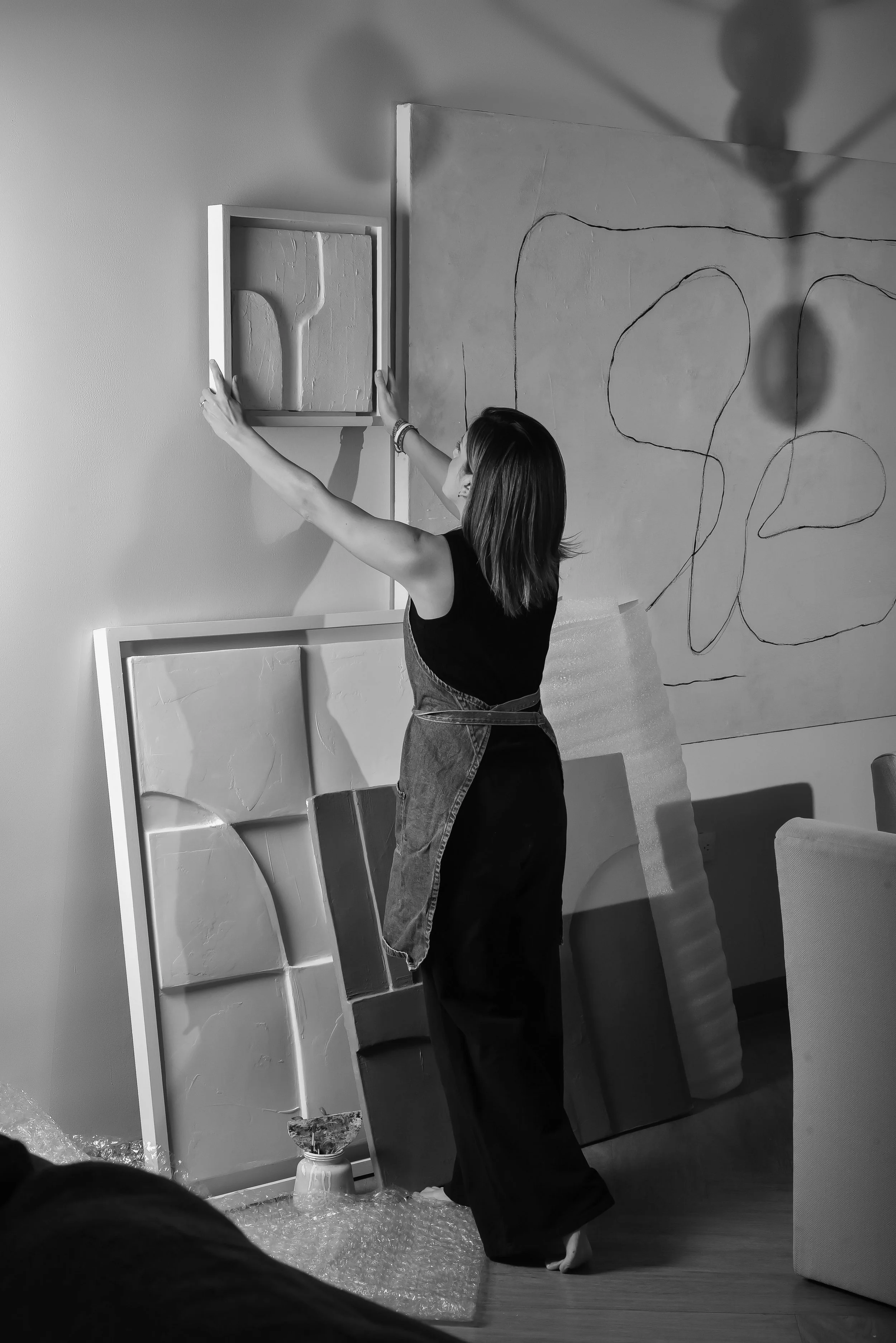 Una mujer organizando obras de arte en una galería o estudio, con varias pinturas en las paredes y en el suelo.