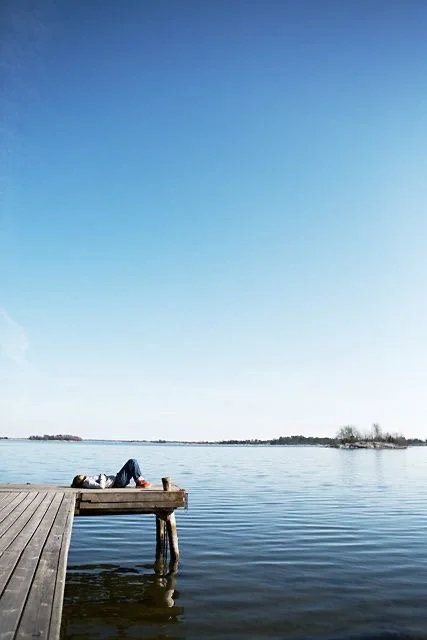 Personne allongée sur une jetée en bois au bord d'un lac calme, sous un ciel bleu clair.