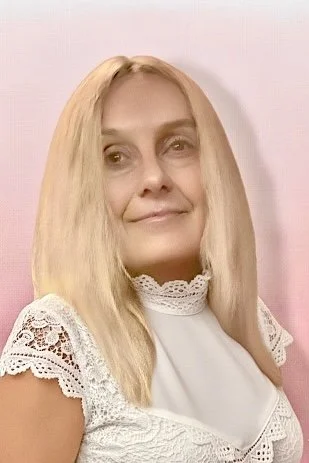 Femme aux cheveux blonds et longs, portant une chemise blanche avec des détails en dentelle, pose devant un fond rose clair.