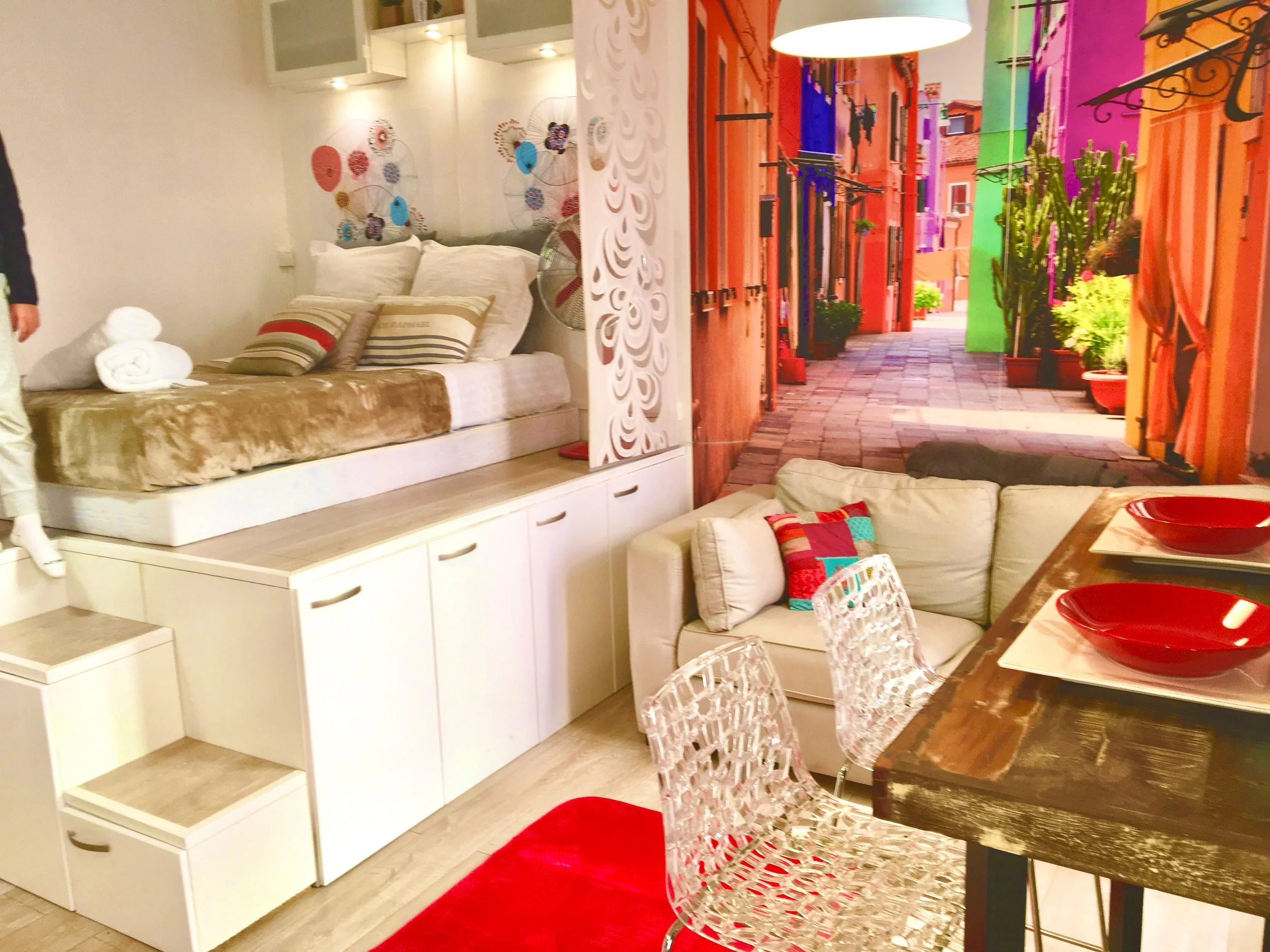 Intérieur d'un appartement avec un coin repas, canapé blanc avec coussins colorés, lit en mezzanine avec des draps beige et des serviettes roulées, et une photo d'une rue colorée en toile de fond