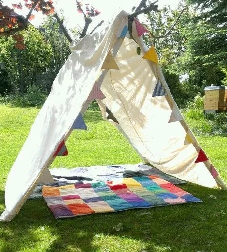 Une tente en tissu blanc décorée de guirlandes de triangles colorés, installée sur une pelouse verte avec un tapis multicolore à l'intérieur.