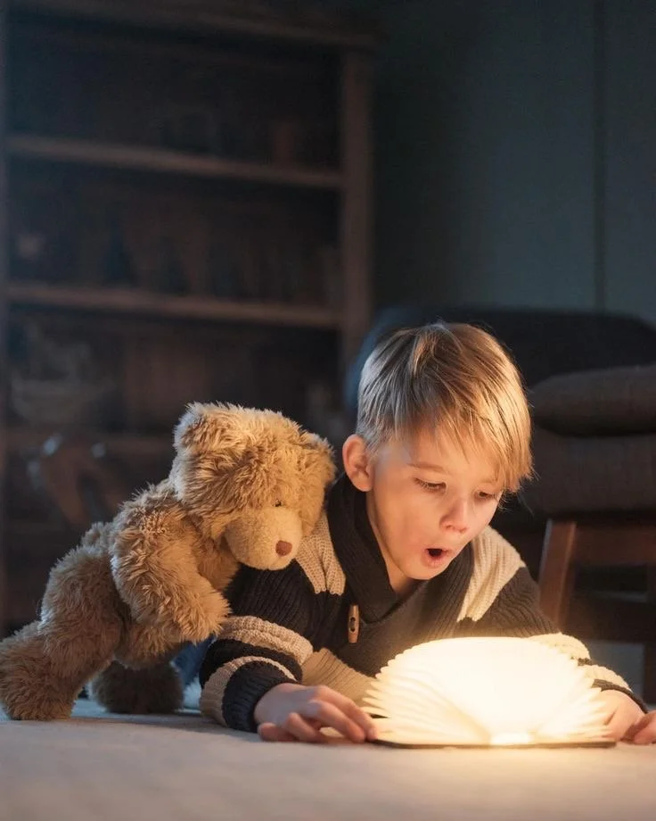 Un jeune garçon lit un livre lumineux à plat sur le sol avec un ours en peluche à ses côtés dans une pièce sombre.