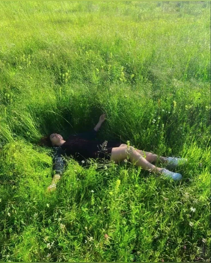 Une femme allongée dans une pelouse verte, tête reposant sur l'herbe, vêtue d'une robe noire, portant des baskets blanches, entourée d'herbes hautes