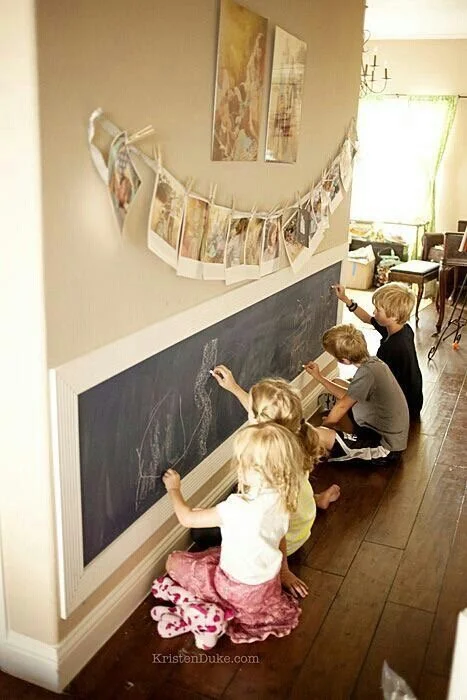 Quatre enfants dessinent avec des craies sur un tableau noir fixé au mur dans une pièce lumineuse.