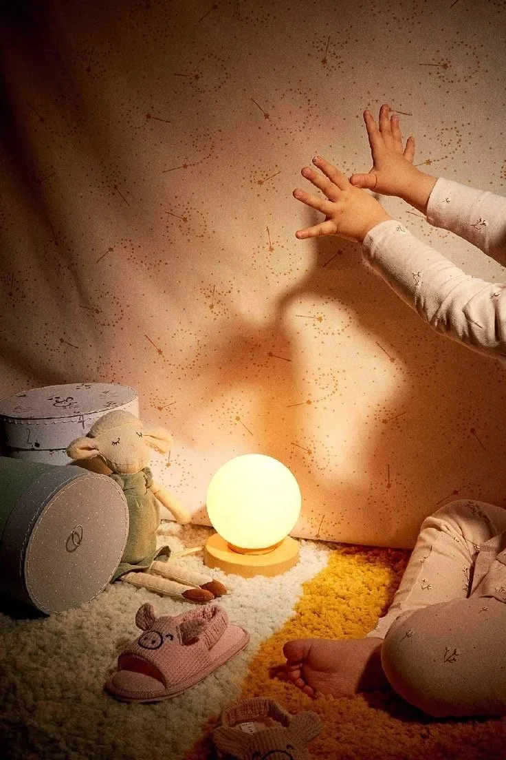 Une scène d'intérieur avec des jouets pour enfants, une lampe ronde allumée, et des mains d'enfant touchant le mur décoratif étoilé.