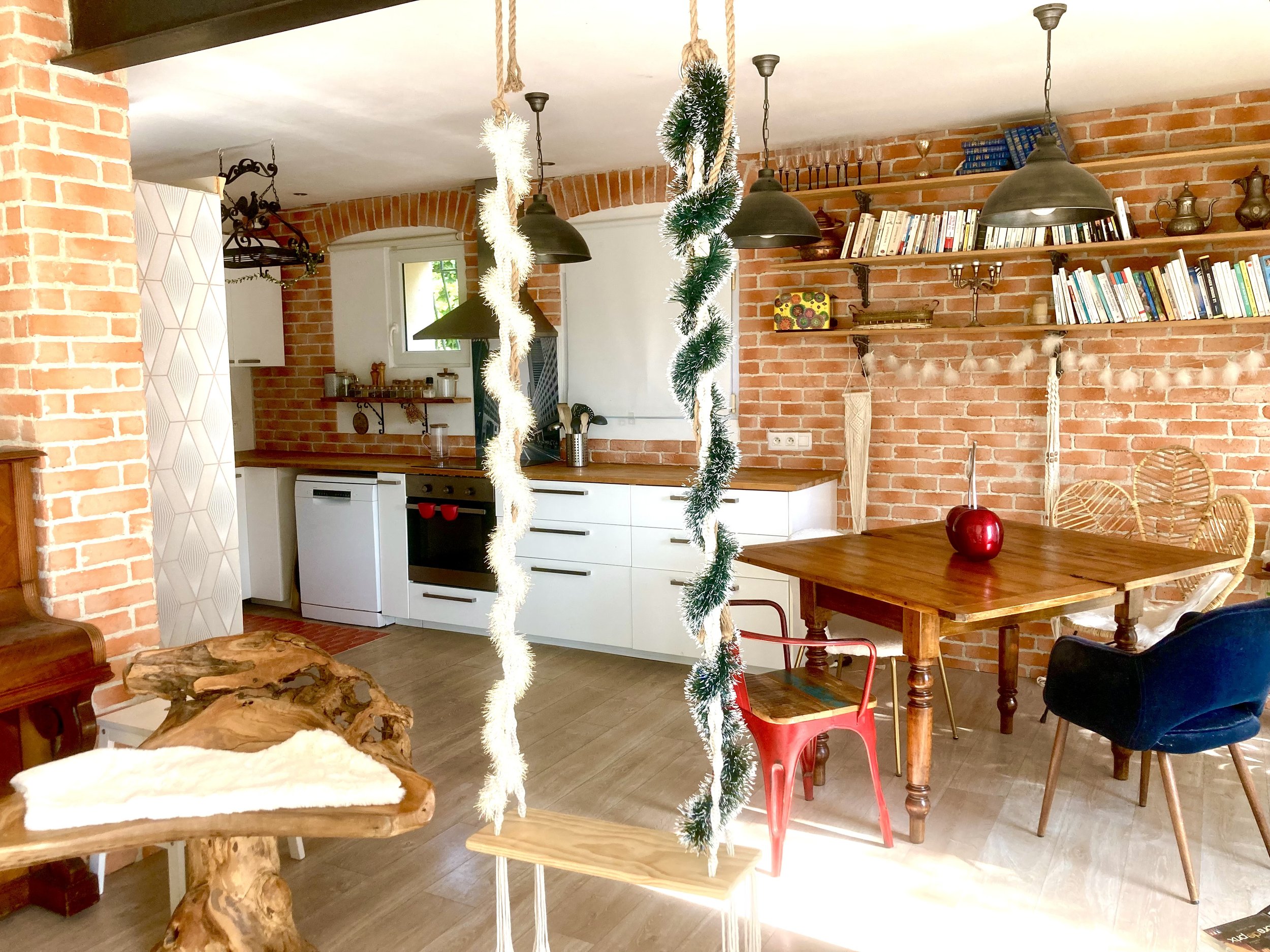 Cuisine ouverte avec murs en briques, table en bois, chaises en rotin et en velours bleu, étagères avec livres et objets décoratifs, guirlande festive, décoration de Noël