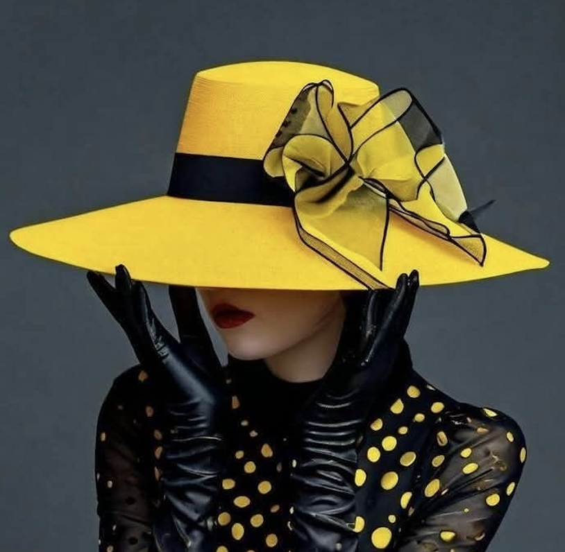 Femme portant un chapeau jaune avec un ruban noir et une décoration en tissu assortie, portant des gants noirs en cuir, habillée d'une robe noire à pois jaunes, posant contre un fond gris.