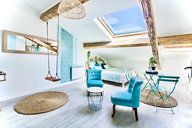 Intérieur d'une chambre sous un toit en pente avec un lit, deux fauteuils bleus, un tapis rond, un hamac, un miroir mural, une table de chevet, et un grand puits de lumière solaire.