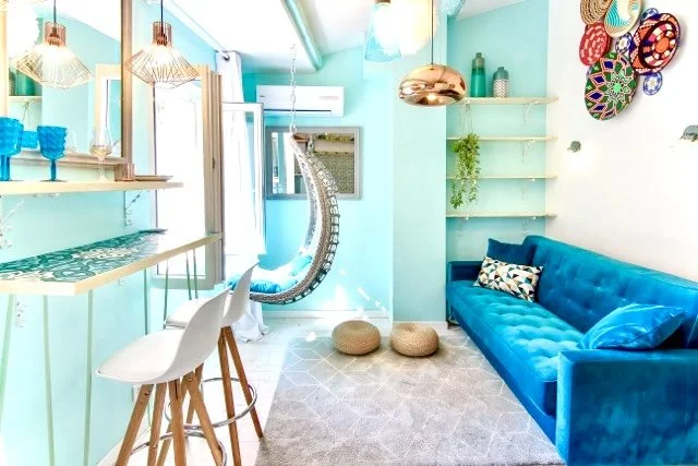 Un salon coloré avec un mur turquoise, un canapé bleu, des étagères flottantes avec des décorations, et un coin lecture avec une balançoire suspendue.