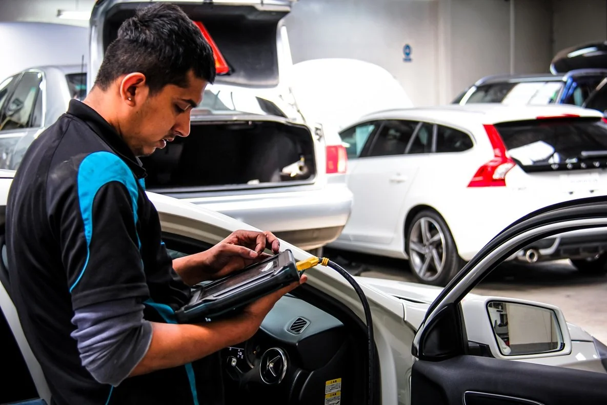 Técnico automotriz revisando un auto con herramienta de diagnóstico en un taller, rodeado de otros vehículos.