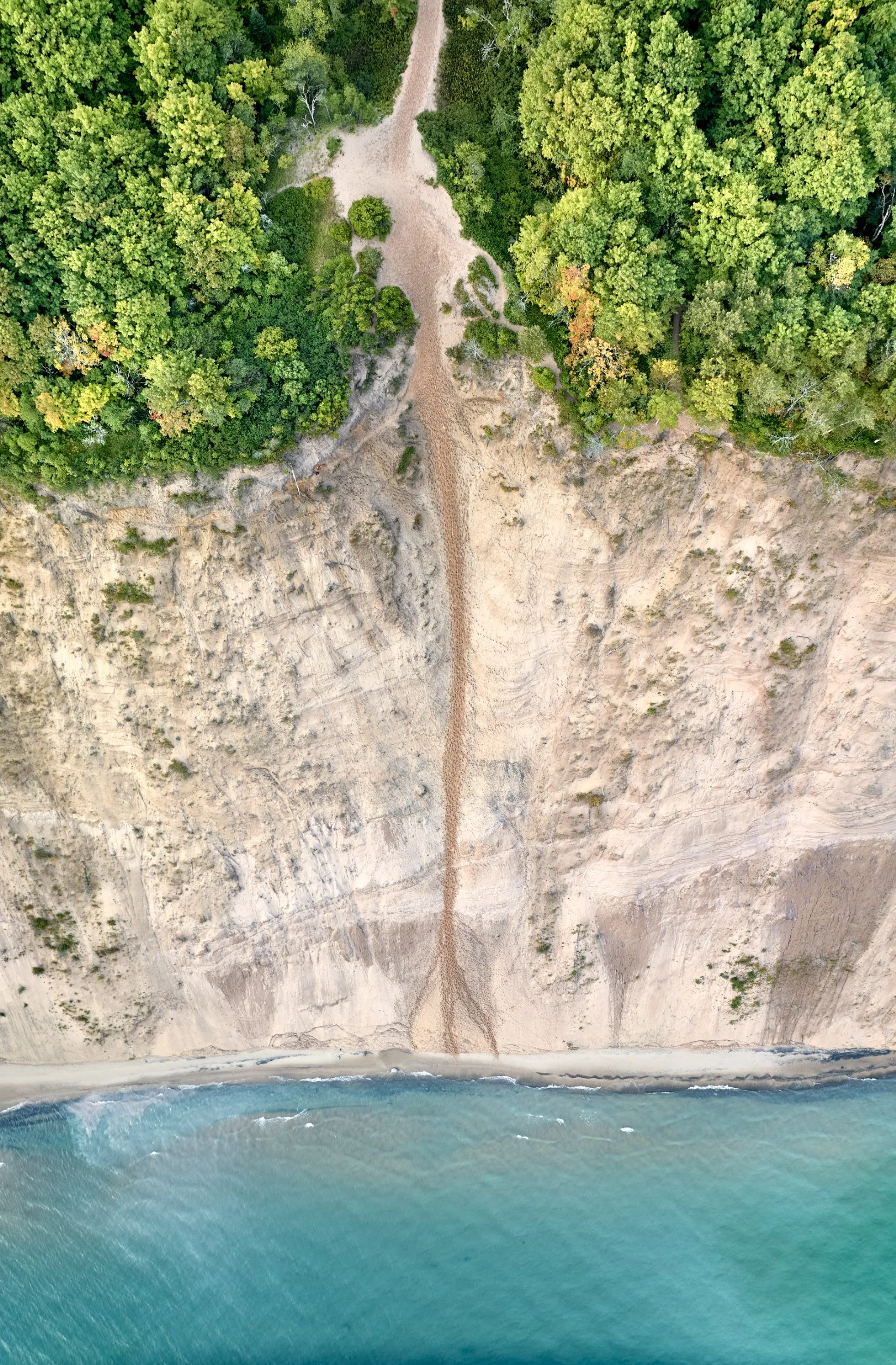An aerial view of an overlook at Pictured Rocks with a blend of green forest, sandy cliff, and the Lake Superior with waves.