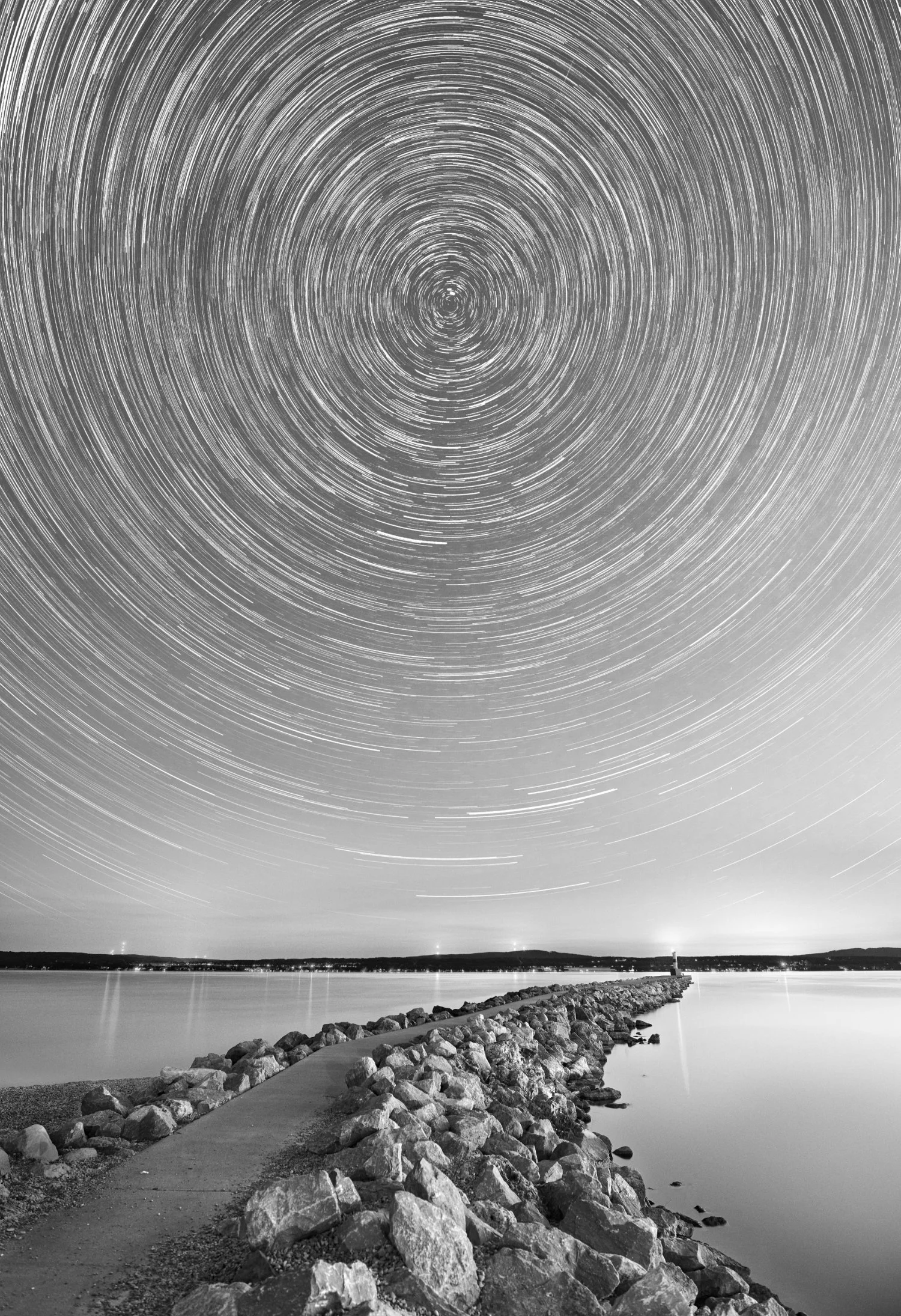 Long exposure black and white photo of star trails in the night sky over a Lake Michigan and the Breakwall in Petoskey, Michigan in this photograph taken by James Manning