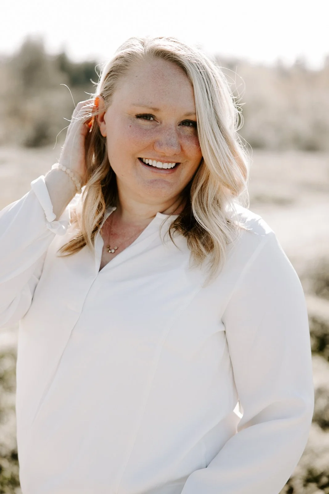 Woman smiling outdoors in natural light, wearing a white blouse, with blurred greenery in the background.