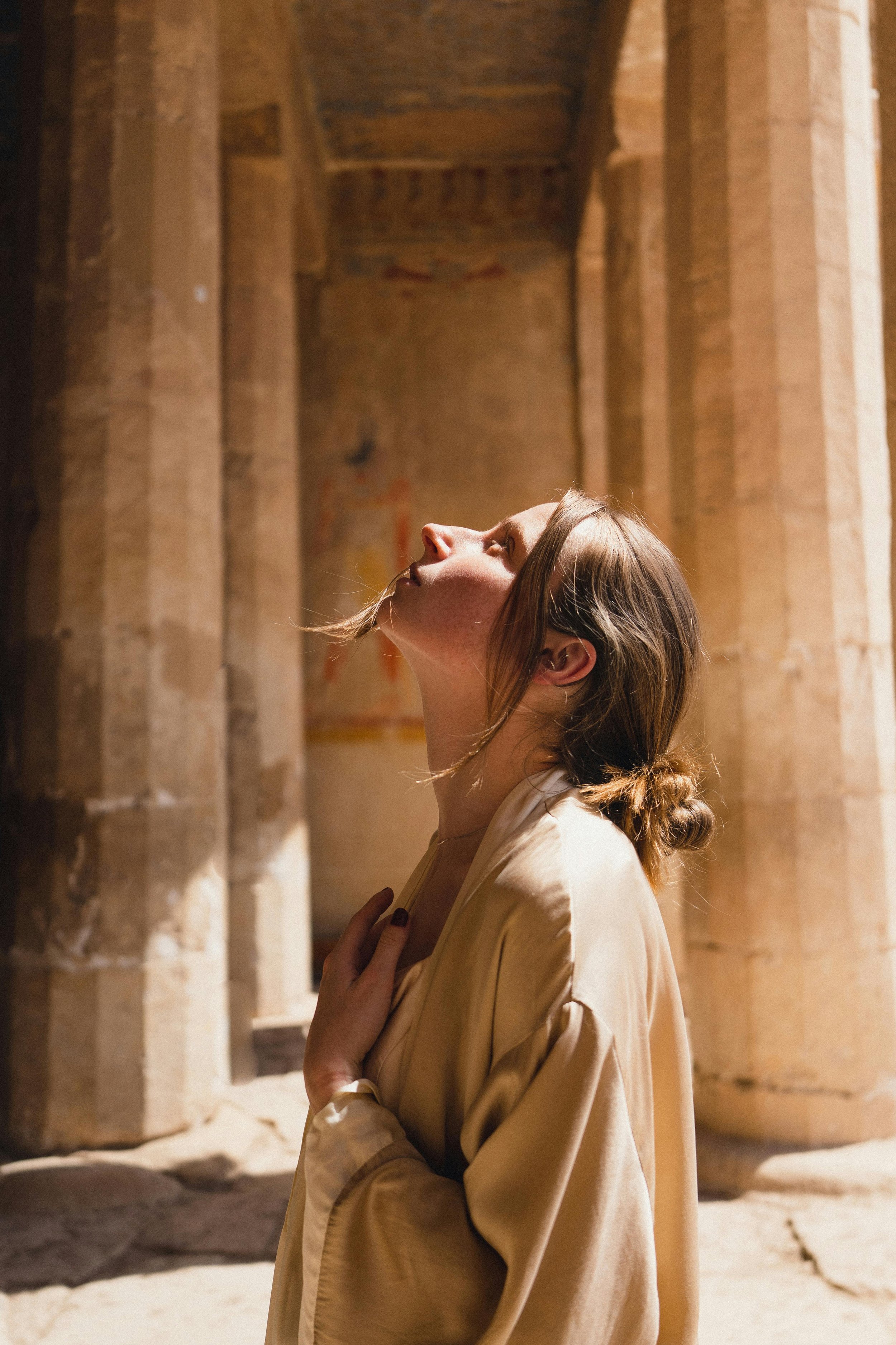 Woman in beige robe looking up in sunlight, standing among ancient stone columns.